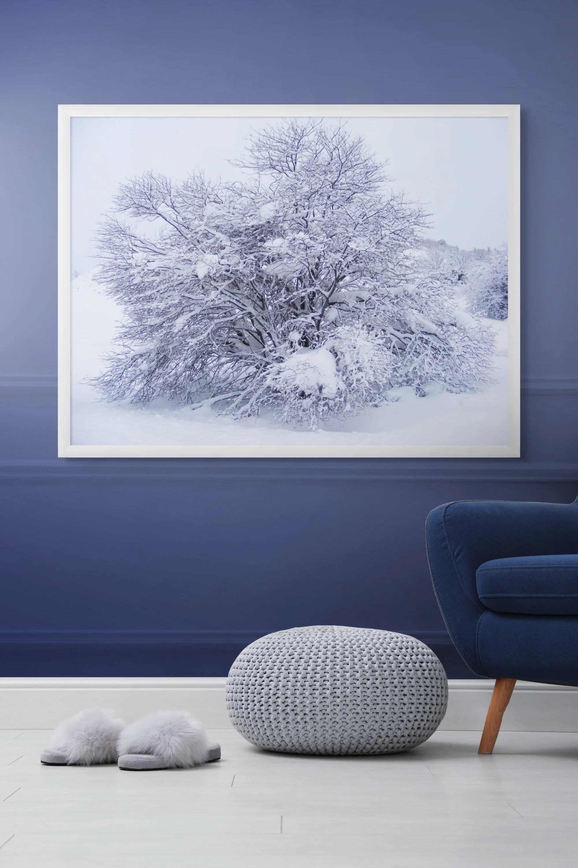 Interior scene with a framed picture of snow-covered trees, a blue armchair, a white knitted ottoman, and fluffy slippers on a white floor against a dark blue wall.