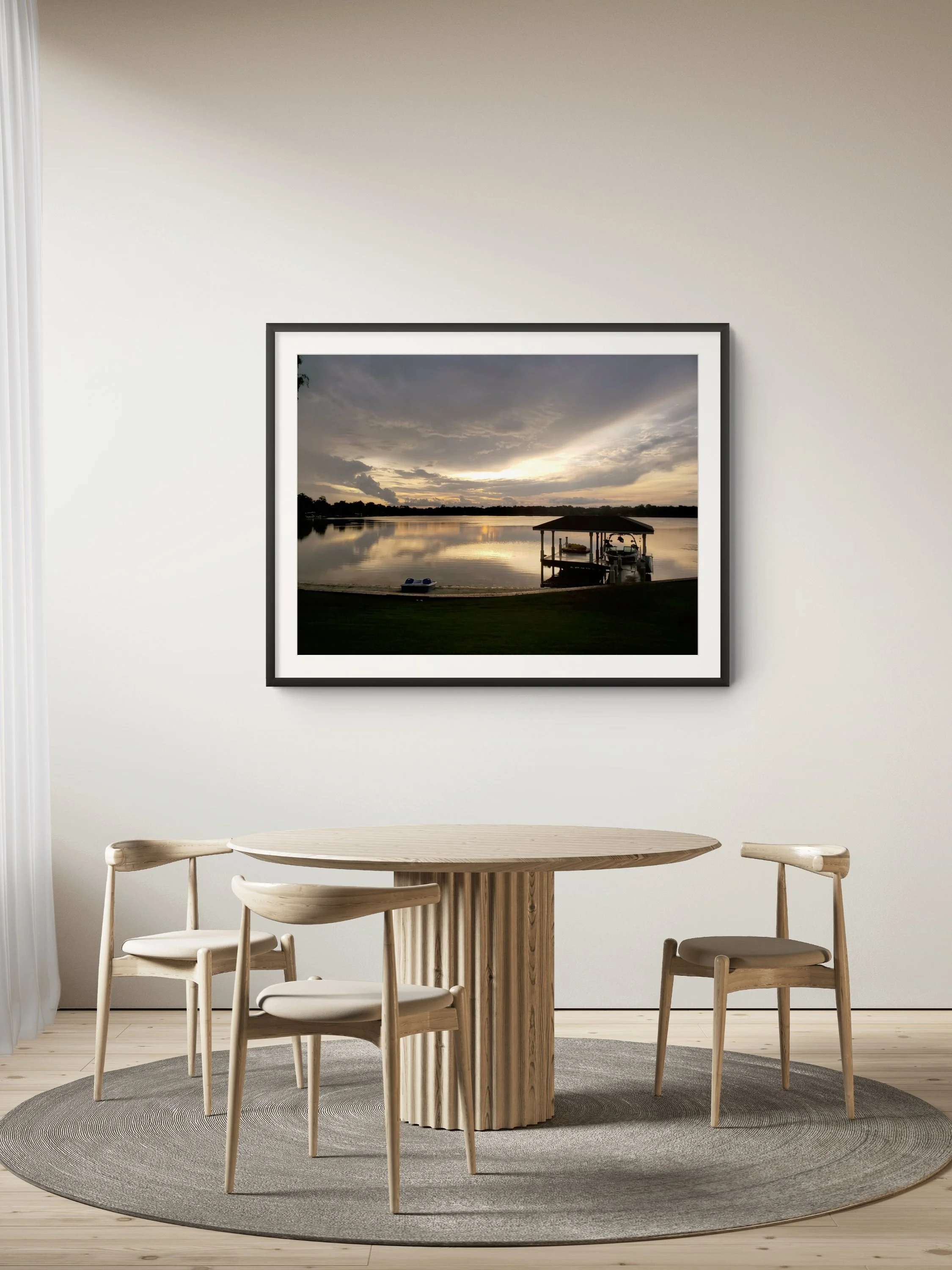 A dining area with a round wooden table, four matching wooden chairs, a gray circular rug, and a large framed landscape photograph of a lake and cloudy sky on the wall.