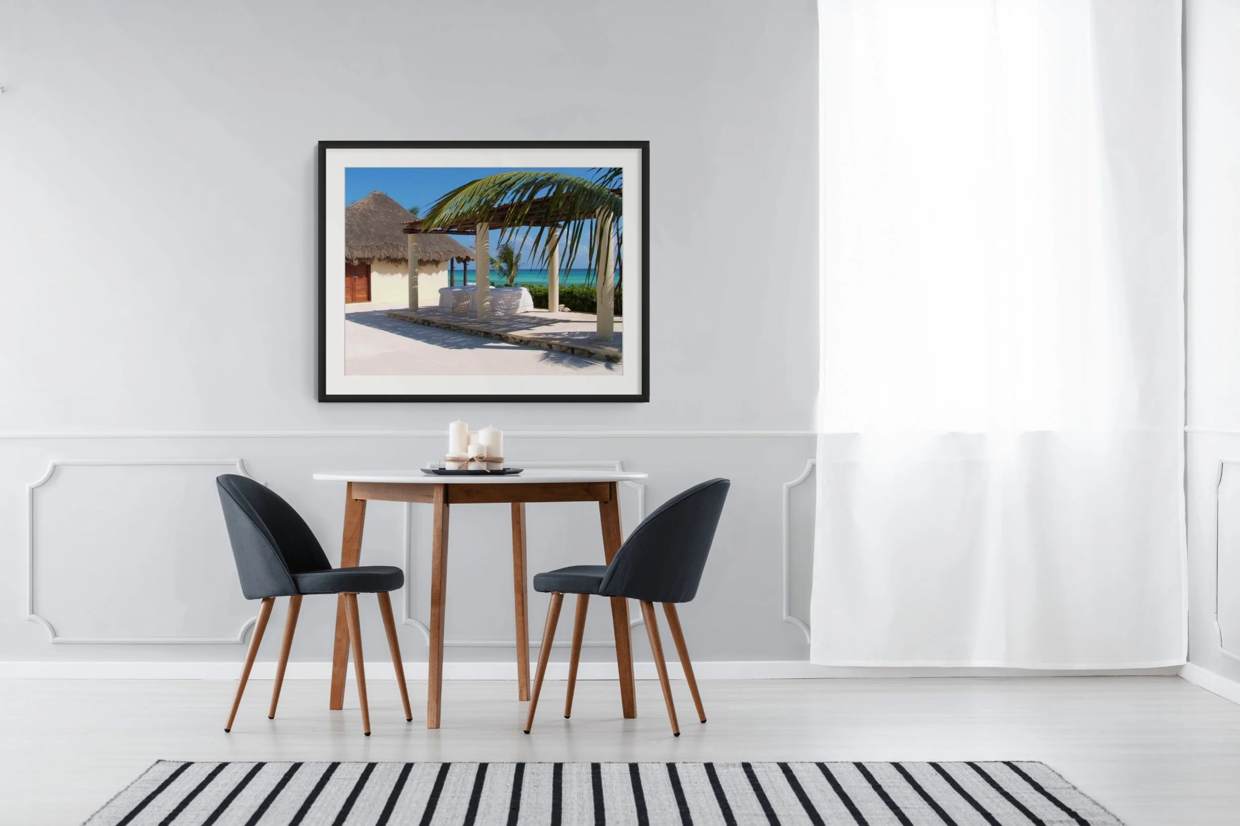 A white wall with a framed tropical beach scene painting, showing a thatched roof hut, palm trees, and the sea, placed above a dining table with four black chairs, a striped rug, and some candles.