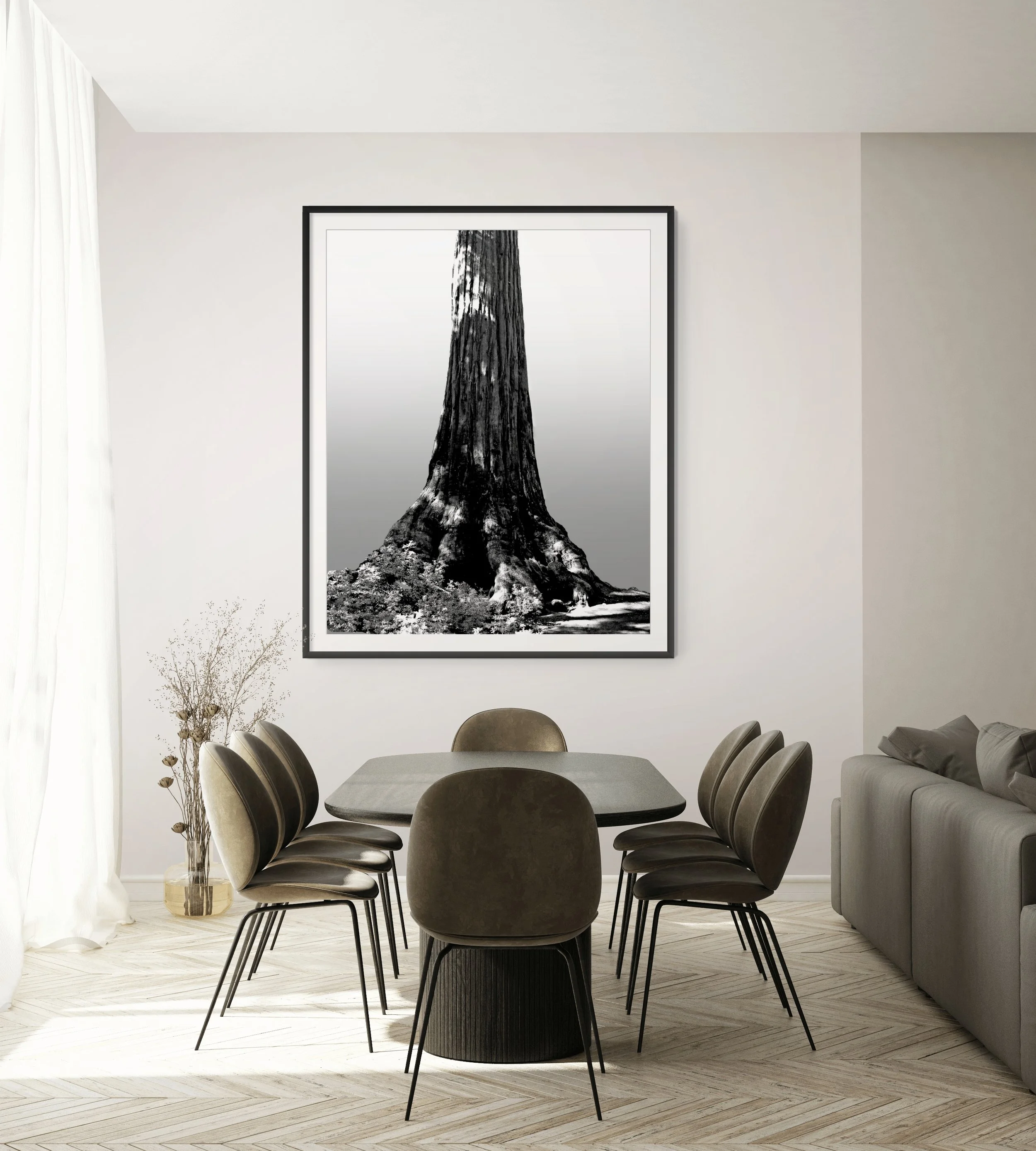 Modern dining room with a black round table, eight black chairs, a gray sofa, a large black and white framed photo of a tree on the white wall, a tall glass vase with bare branches, light wood flooring, and white curtains.