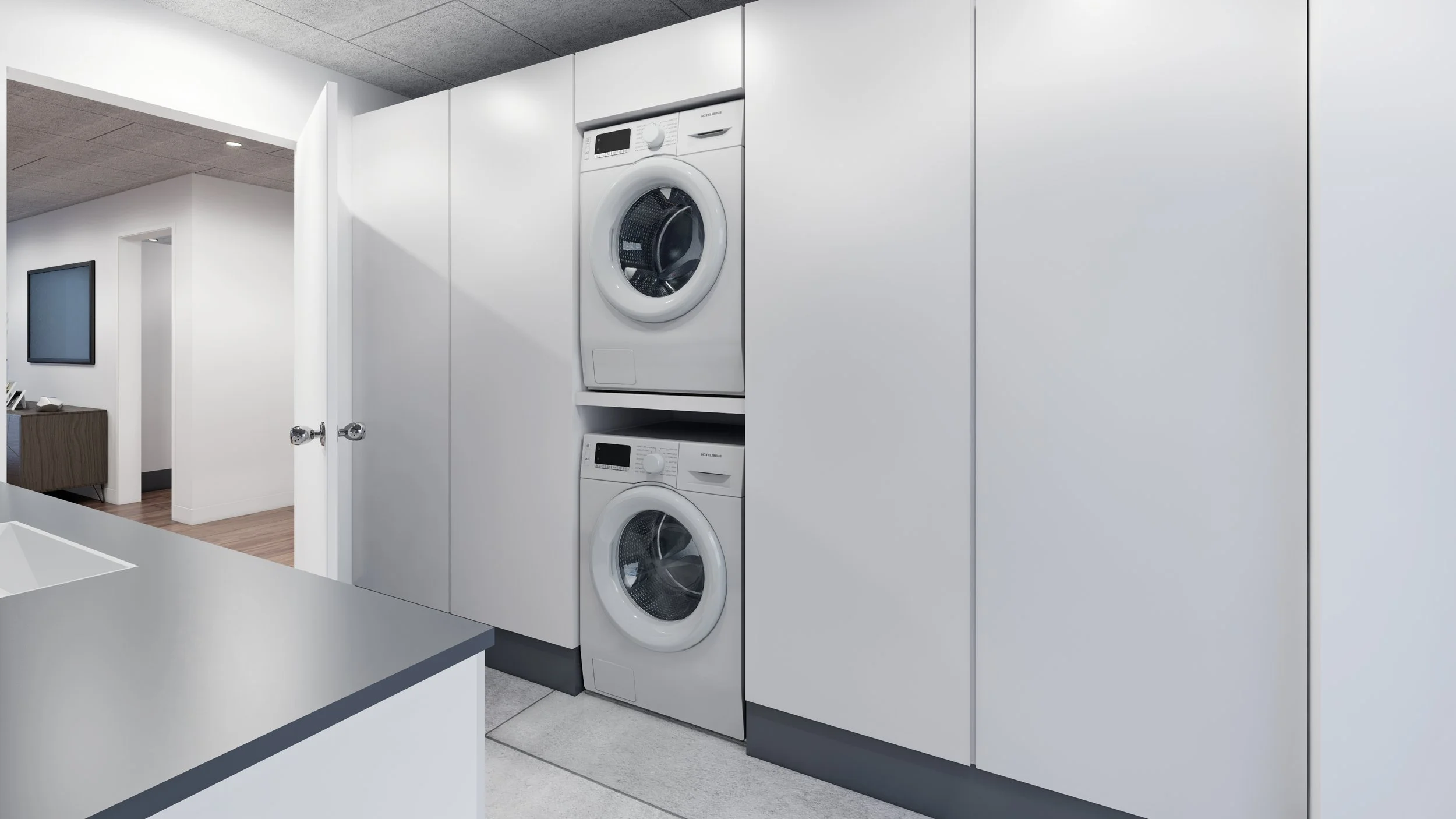 Beskrivelse af billedet: Der er en vaskemaskine og tørretumbler stablet i et skabsvægpanel i et vaskerum. Vaskemaskinerne er hvide, og væggen og skabene er også hvide. Der er en adgangsvej til et opholdsrum med trægulv og en væg med et fladskærms-tv 