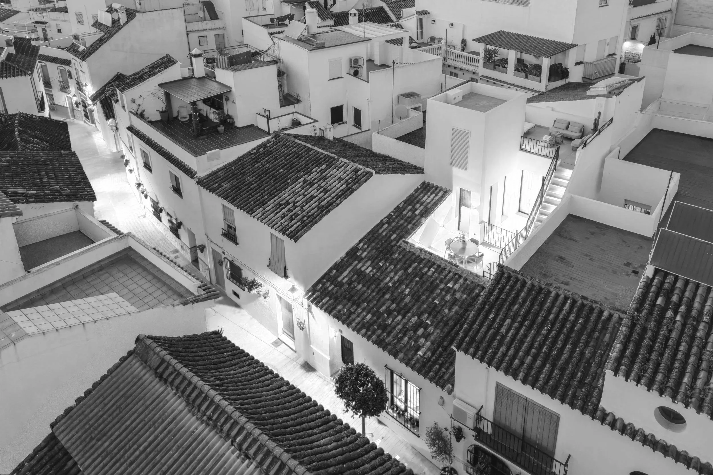 Black and white aerial view of white Mediterranean-style buildings with tiled roofs, some featuring balconies, terraces, and outdoor furniture.