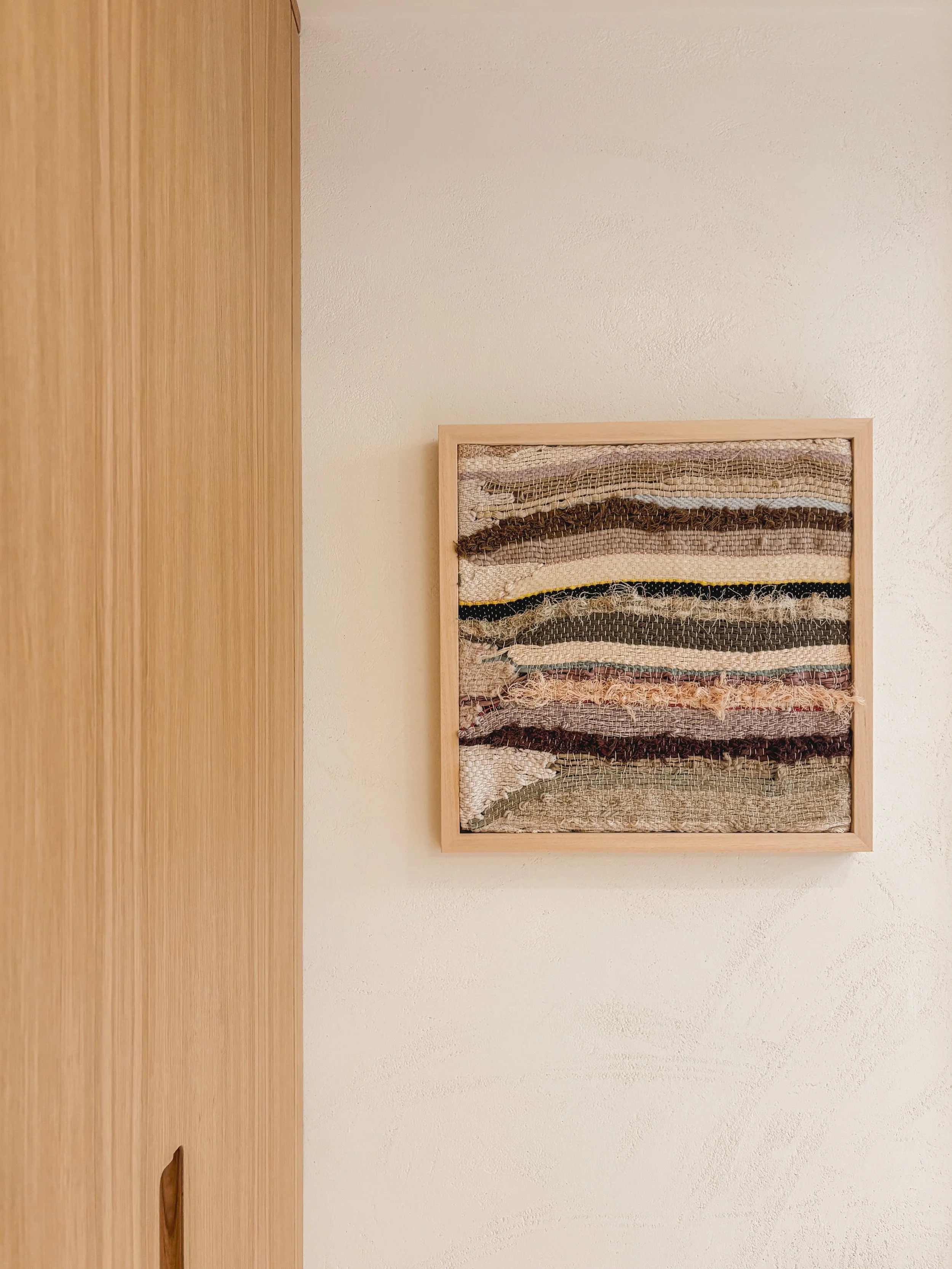 A framed wall art piece made of layered fabric strips in shades of brown, beige, cream, and black, hanging on a light-colored wall next to a wooden door.