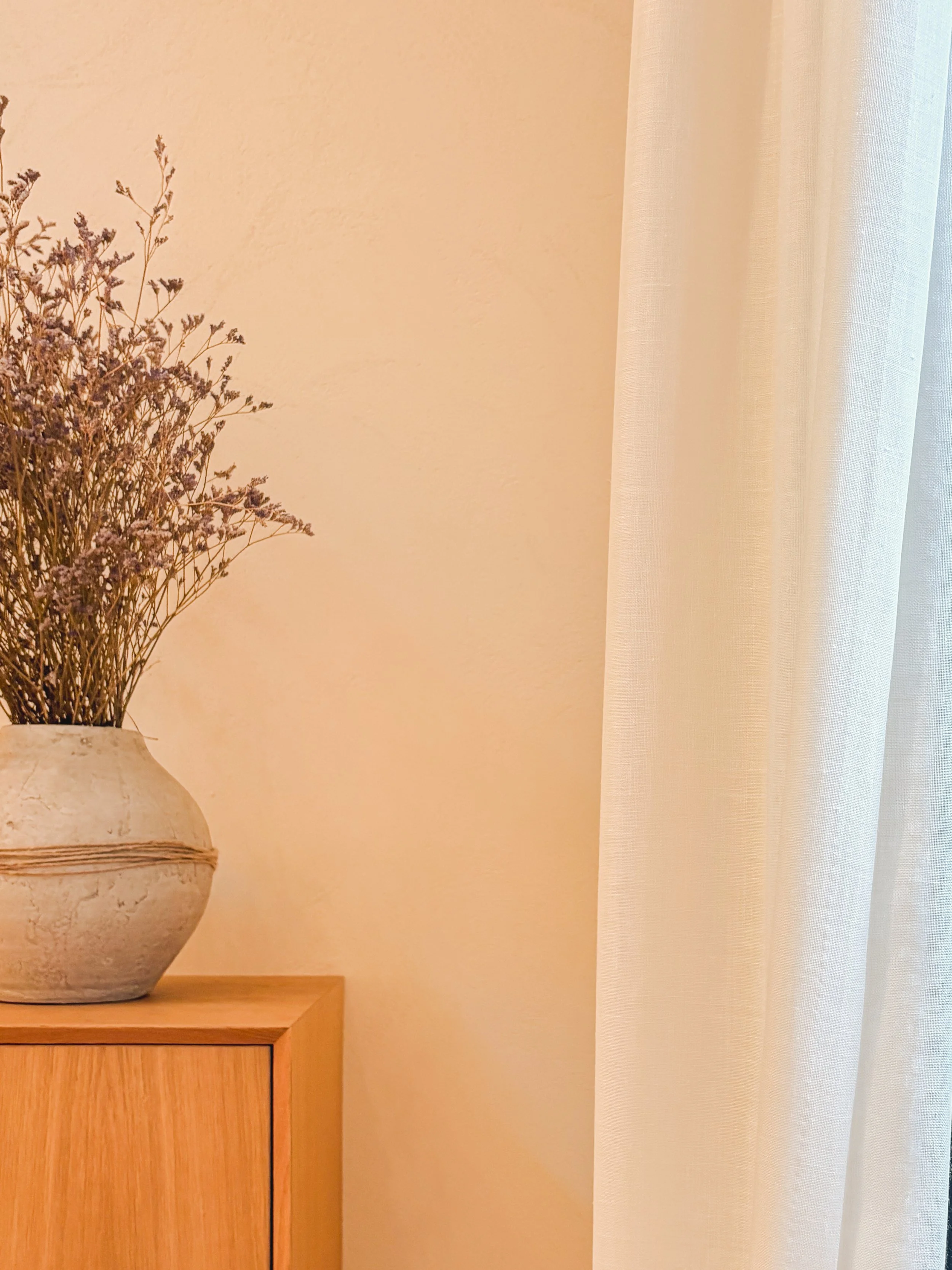 A beige vase with dried purple flowers on a wooden surface next to a white curtain and a light-colored wall.