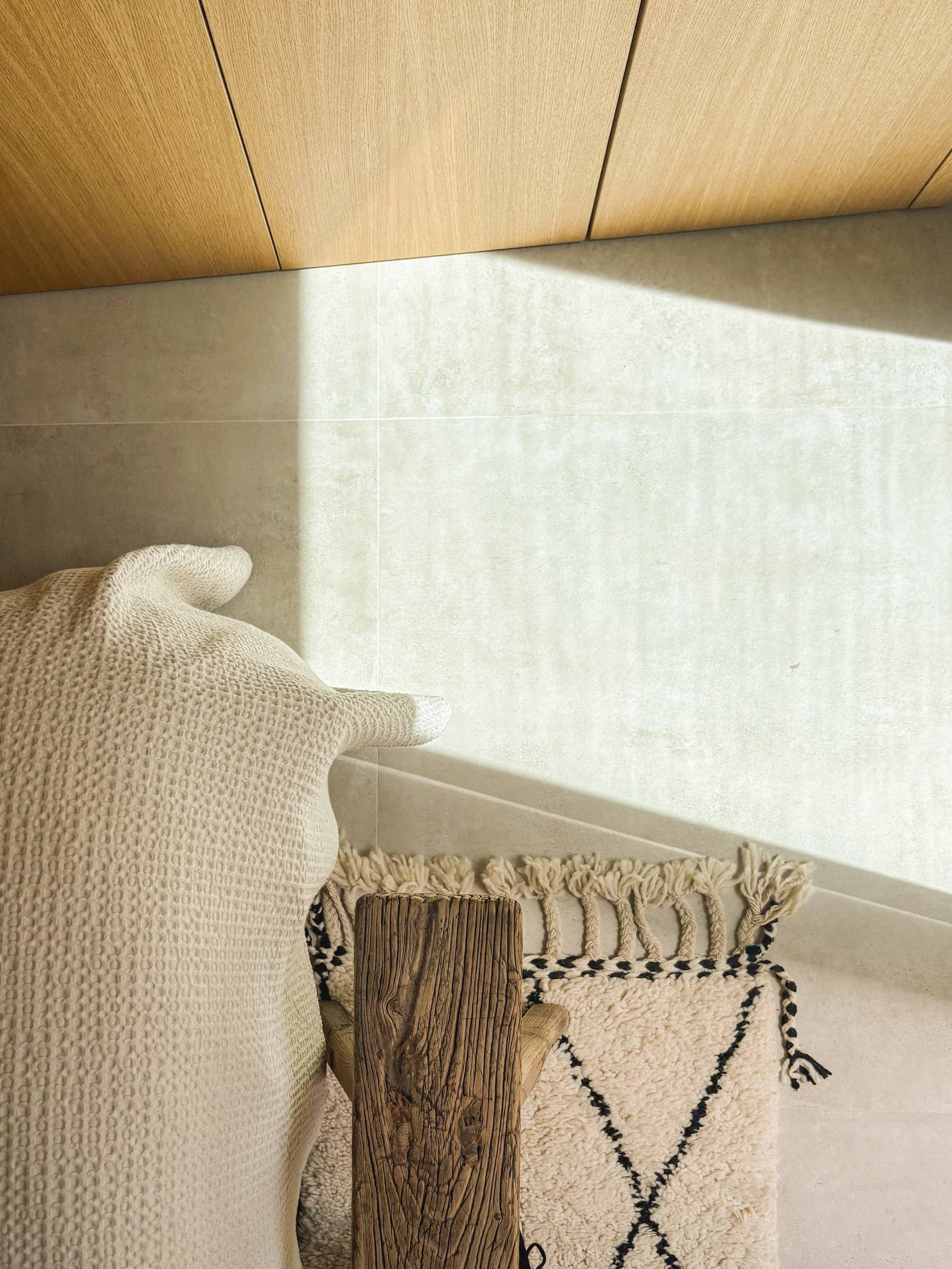 Part of a beige textured fabric armchair, a wooden furniture piece, a patterned rug, and a corner of a light-colored tiled floor with a wooden cabinet and a shadow cast on the floor.