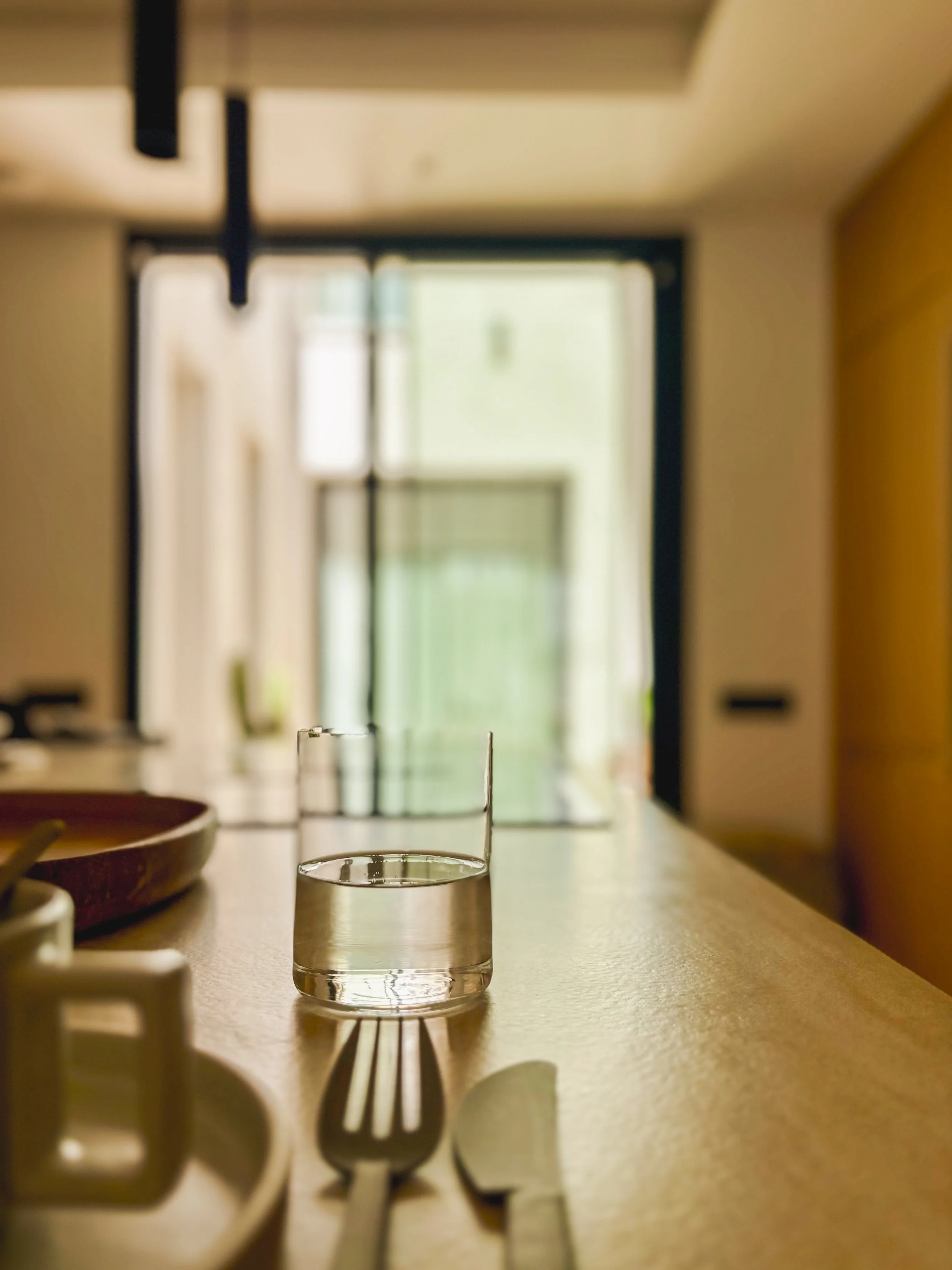 A glass of water, a fork, and a spoon on a wooden counter in a kitchen with a window in the background.