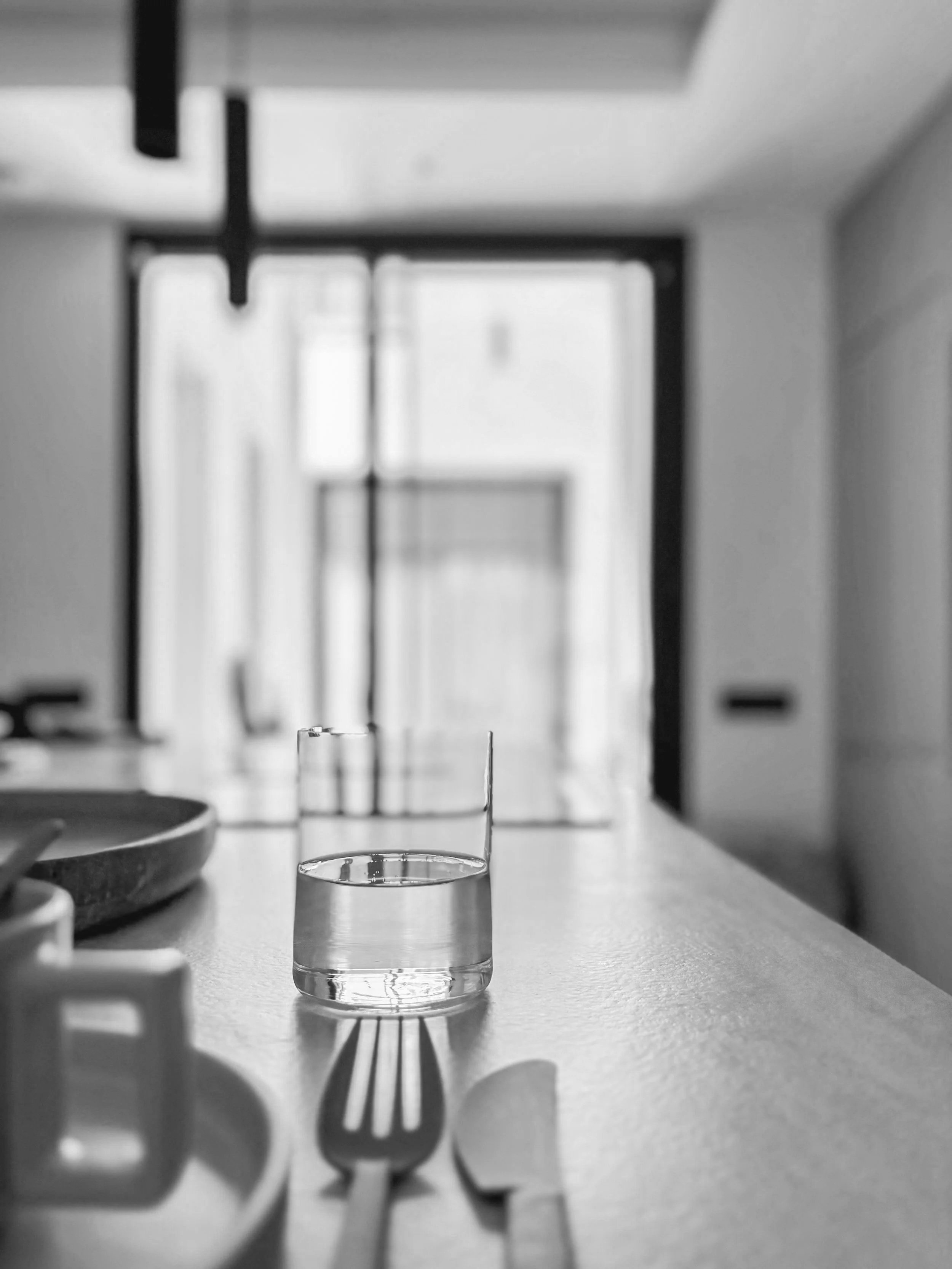 A glass of water on a kitchen counter with a fork and knife in front, and a mug partially visible on the left, in black and white.