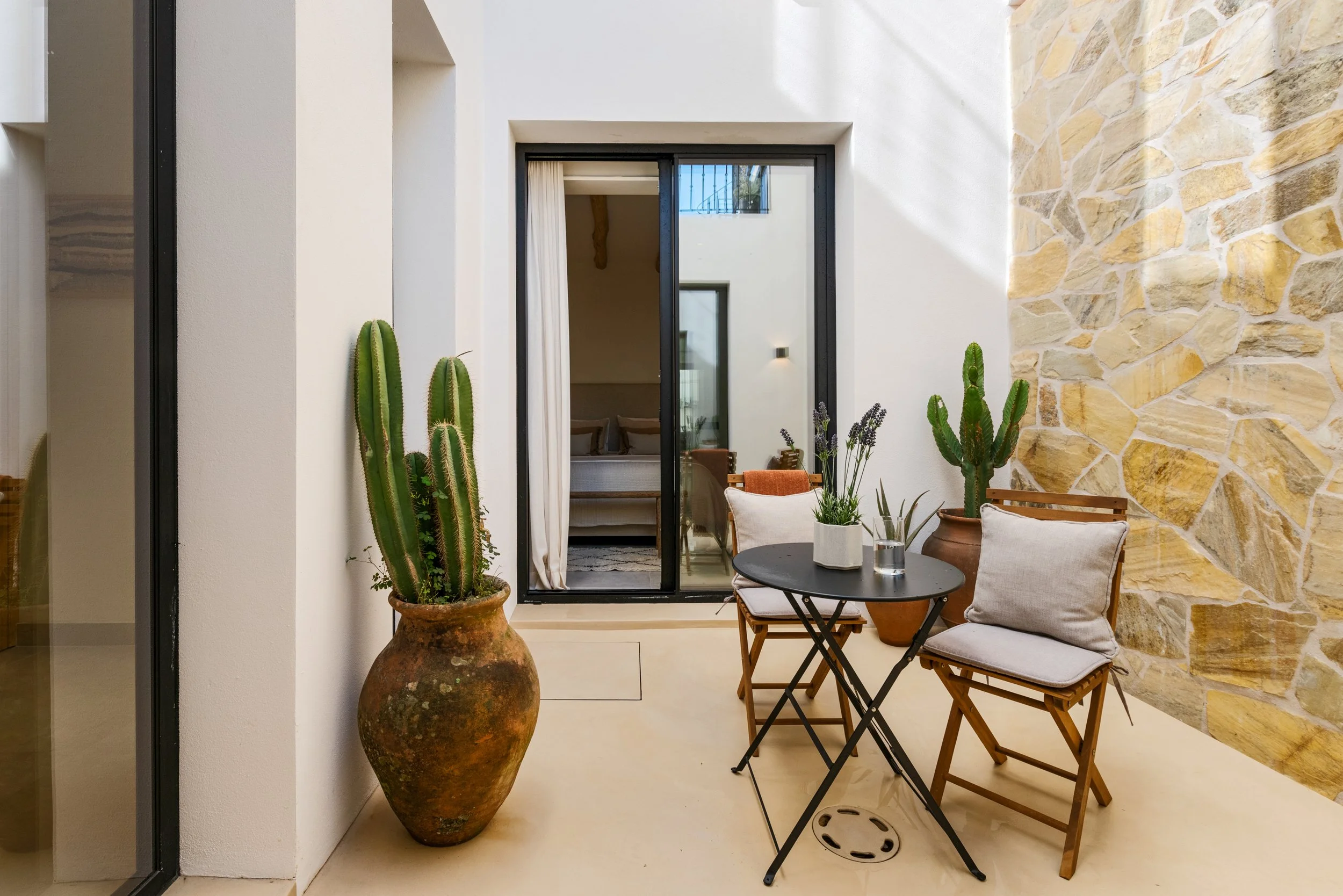 Small outdoor patio with a round black table, two wooden chairs with cushions, potted cacti and lavender, and a sliding glass door leading to an indoor living room.
