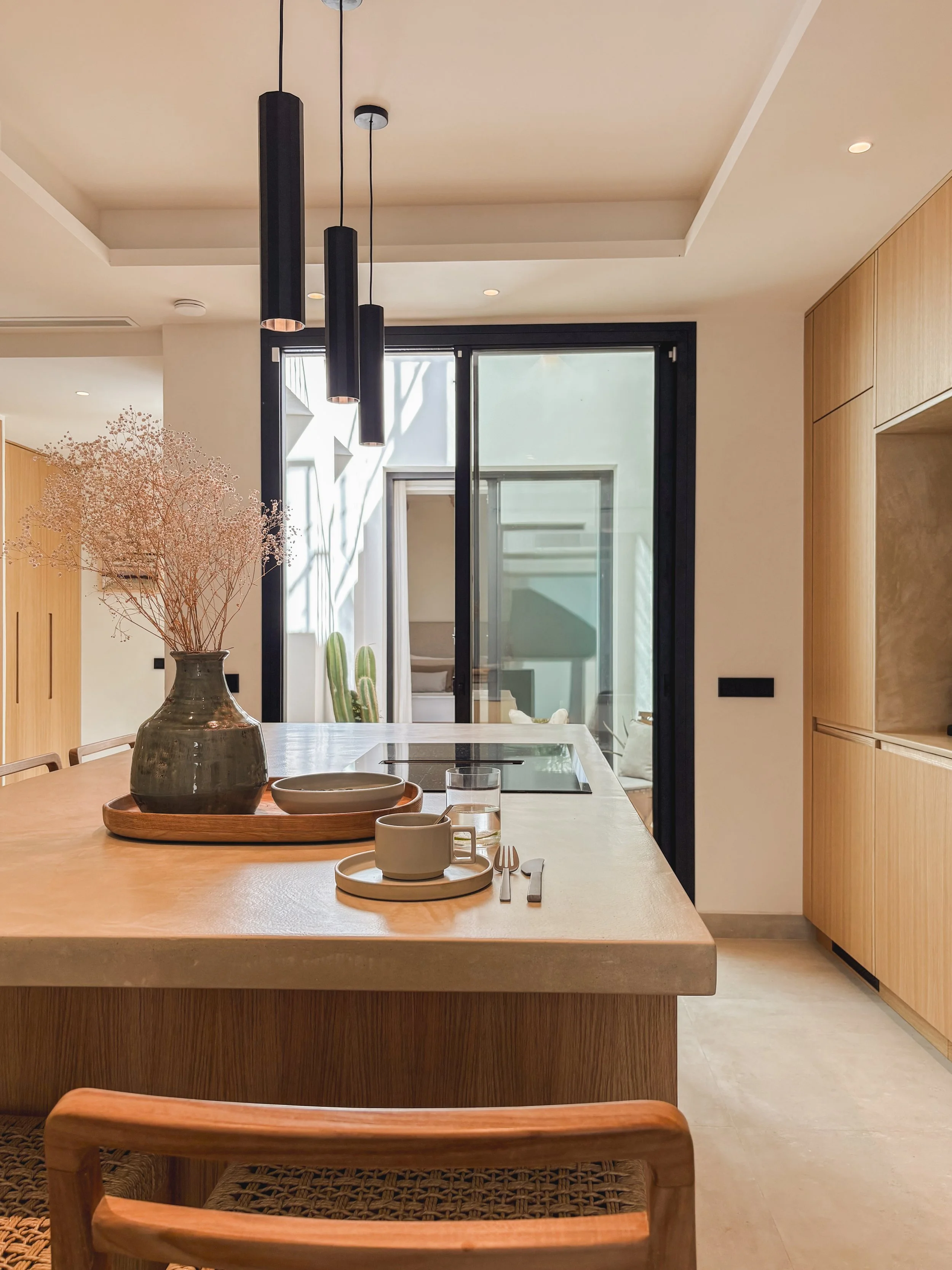 A modern kitchen with a wooden countertop, black pendant lights, and a sliding glass door leading outside, decorated with a vase of dried flowers and tableware.