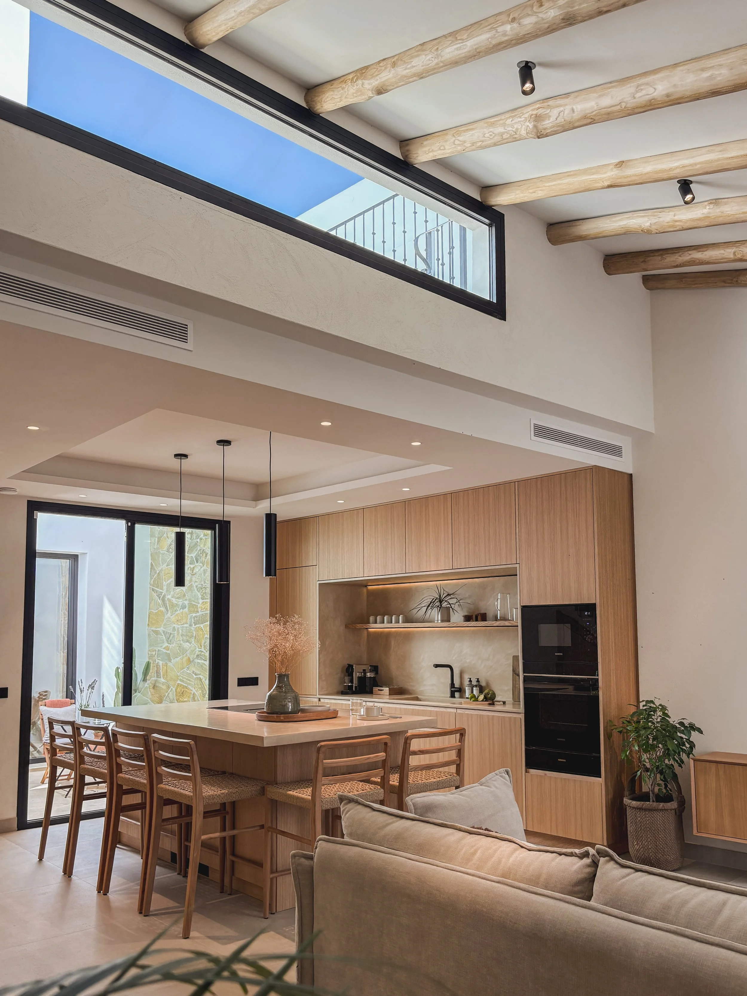 Interior of a modern kitchen and living room with natural wood cabinetry, a kitchen island with bar stools, a beige couch, large windows letting in natural light, and exposed wooden ceiling beams.