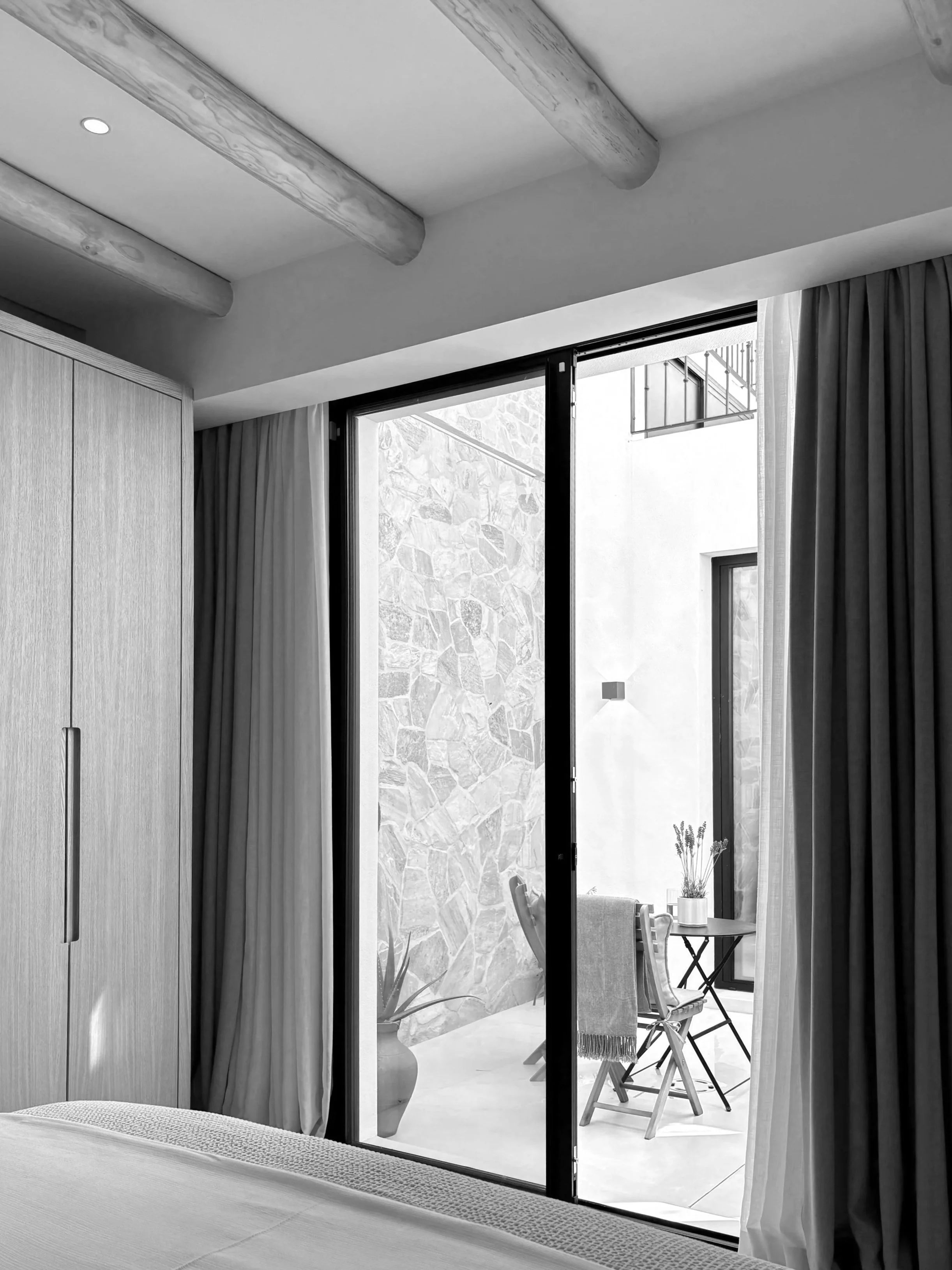 Interior view of a bedroom with a sliding glass door leading to an outdoor patio with a small table and chairs, plant pot, and stone wall.