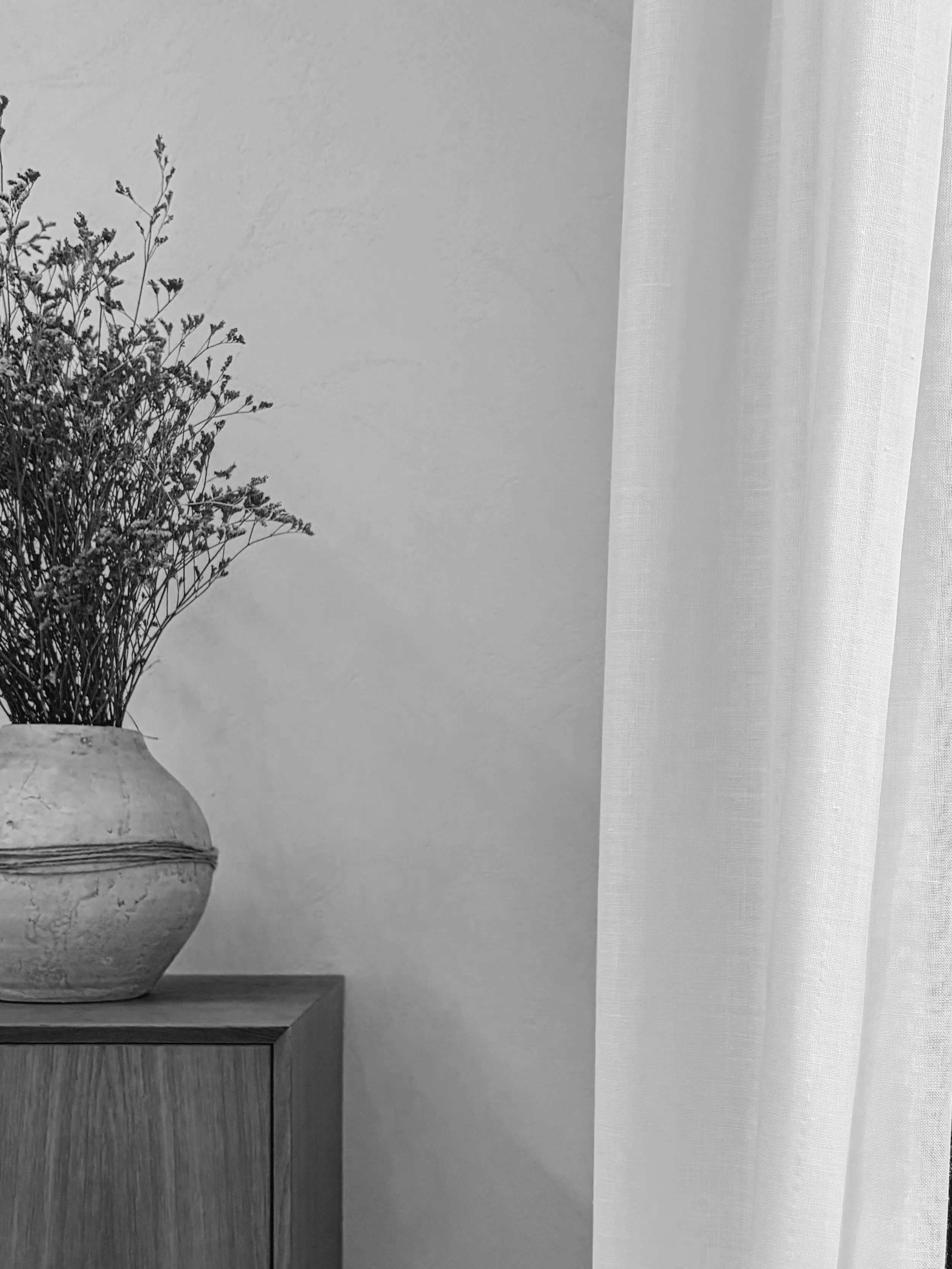 A round, textured ceramic vase holding dried flowers, placed on a wooden surface next to a sheer curtain on the right.