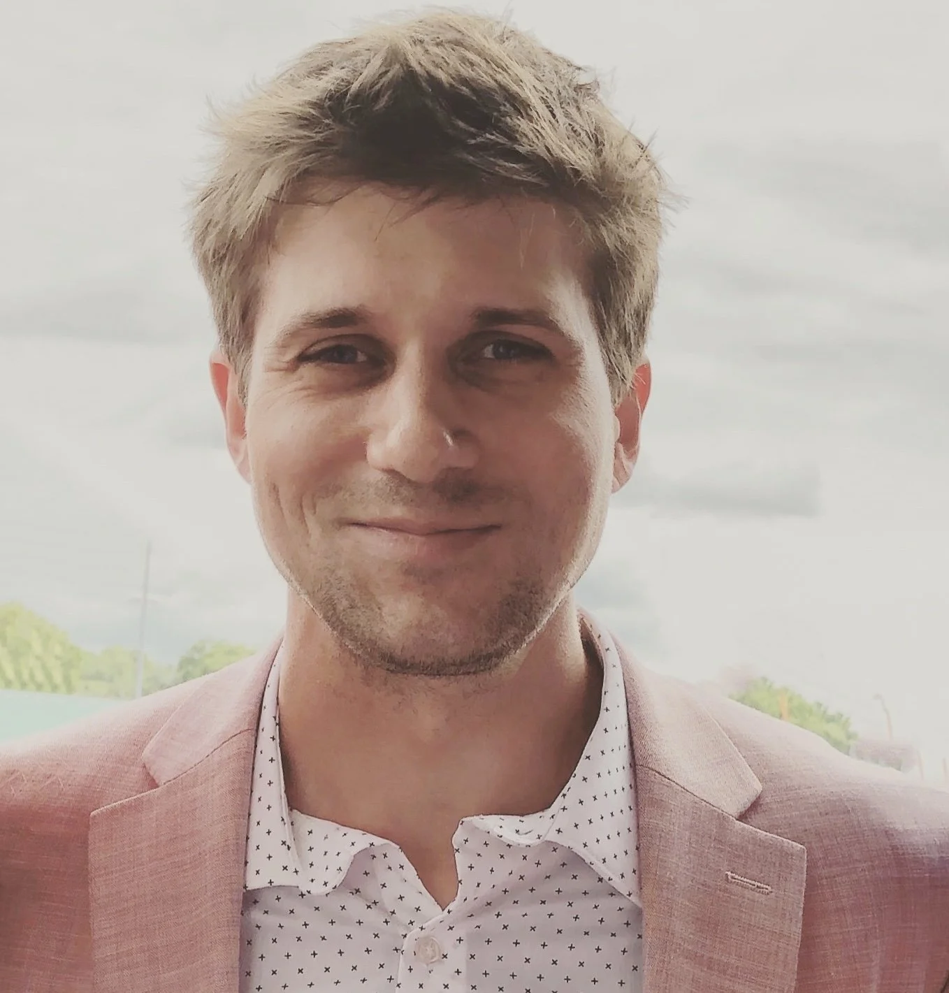 Portrait of a young man with light brown hair, smiling, wearing a pink blazer and a white shirt with small black dots, outdoors on a cloudy day.