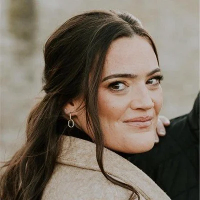 Close-up of a woman with long brown hair, wearing earrings and a beige coat, smiling softly at the camera.