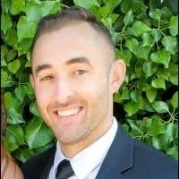 A man smiling in front of green leafy plants, dressed in a formal suit and tie.