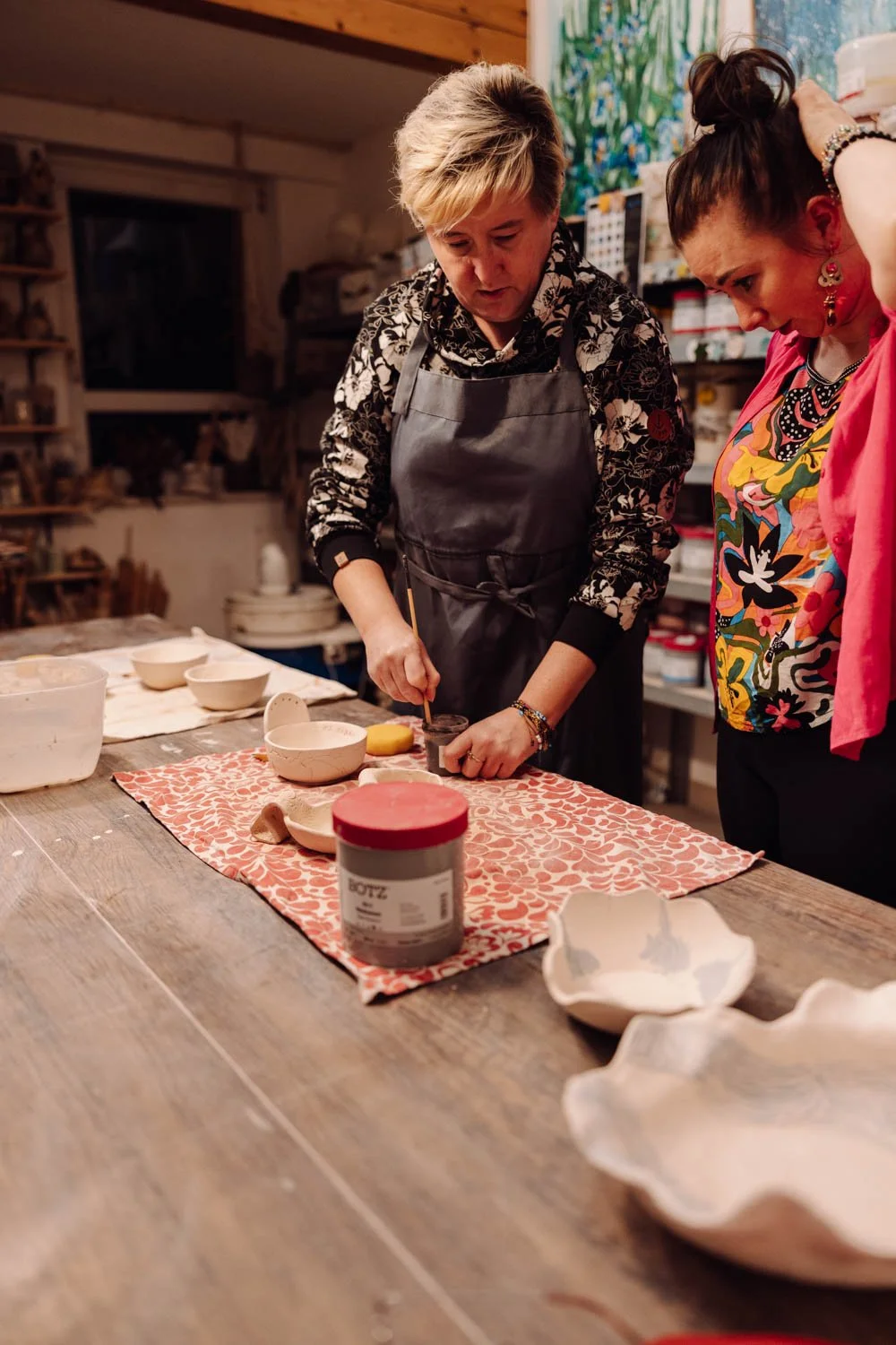 Two women working on pottery or ceramics in a cozy, well-lit studio with shelves of supplies in the background.