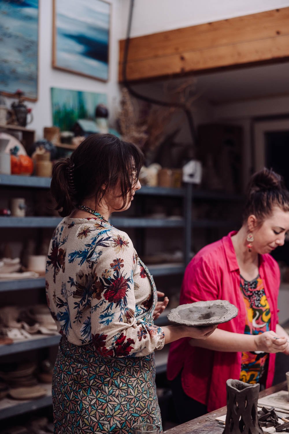 Two women in a pottery studio, one holding a pottery piece and the other examining a clay project, surrounded by shelves filled with pottery and artwork.