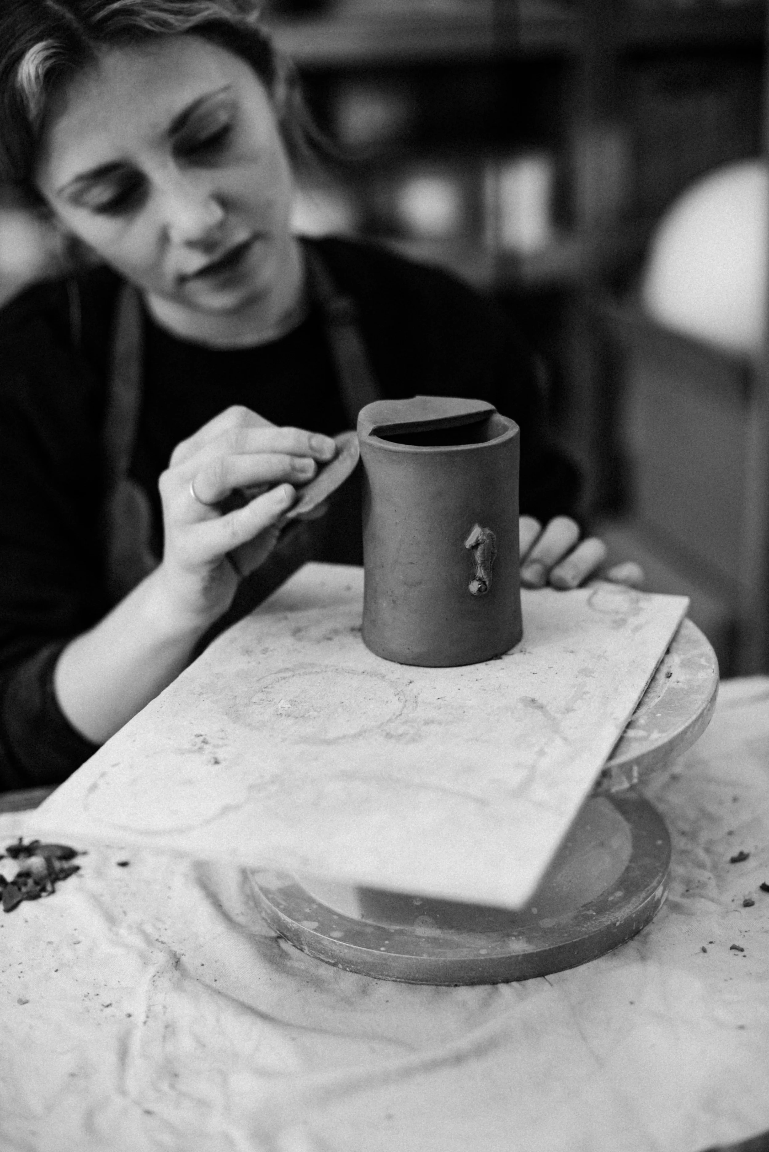 A woman is shaping a ceramic object in a pottery studio, with tools and pottery on the table.