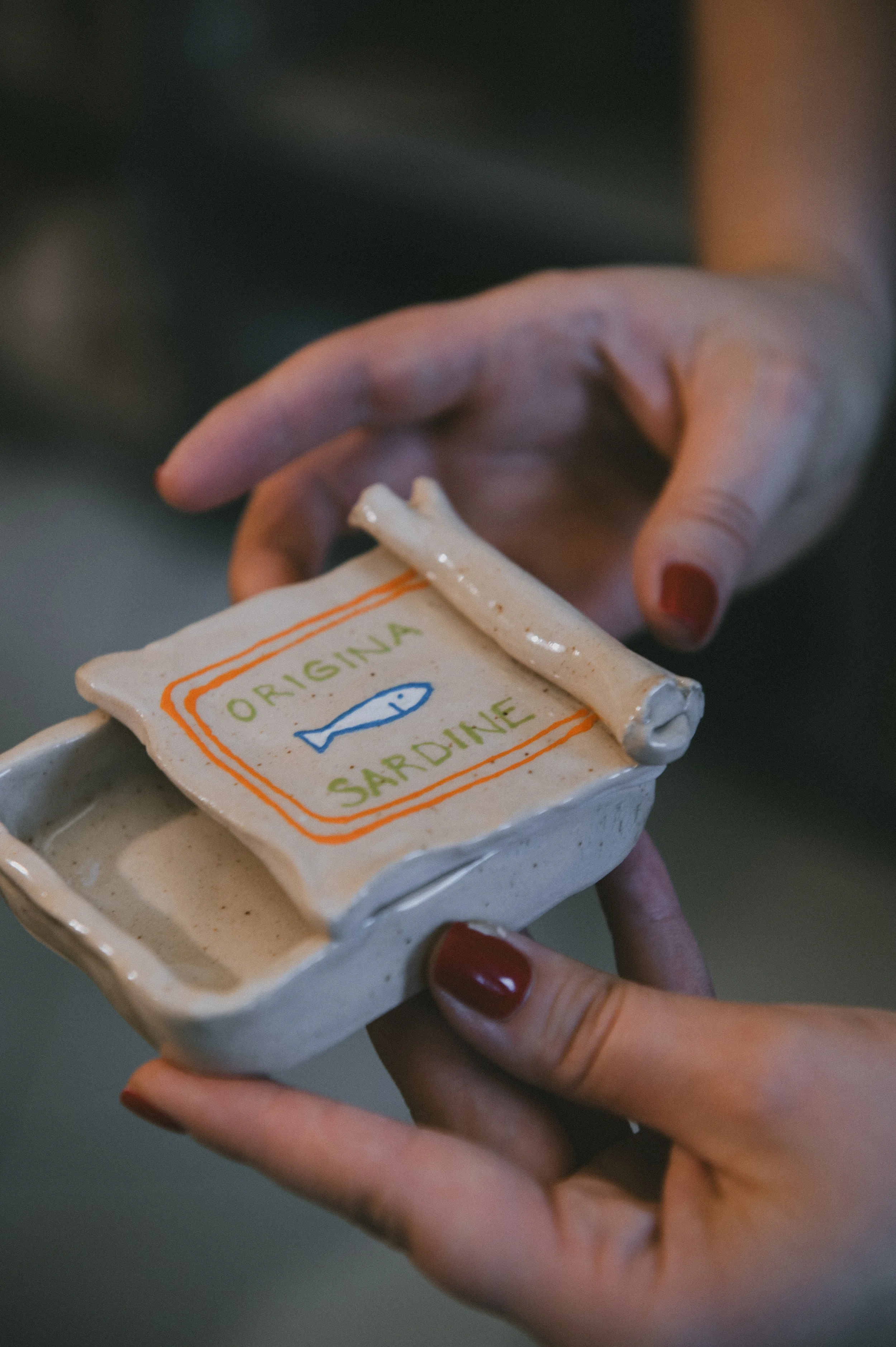 A person holding a small ceramic dish labeled "Original Sardine" with a hand-drawn blue fish and a rolled piece of ceramic resembling a fish skeleton on top.