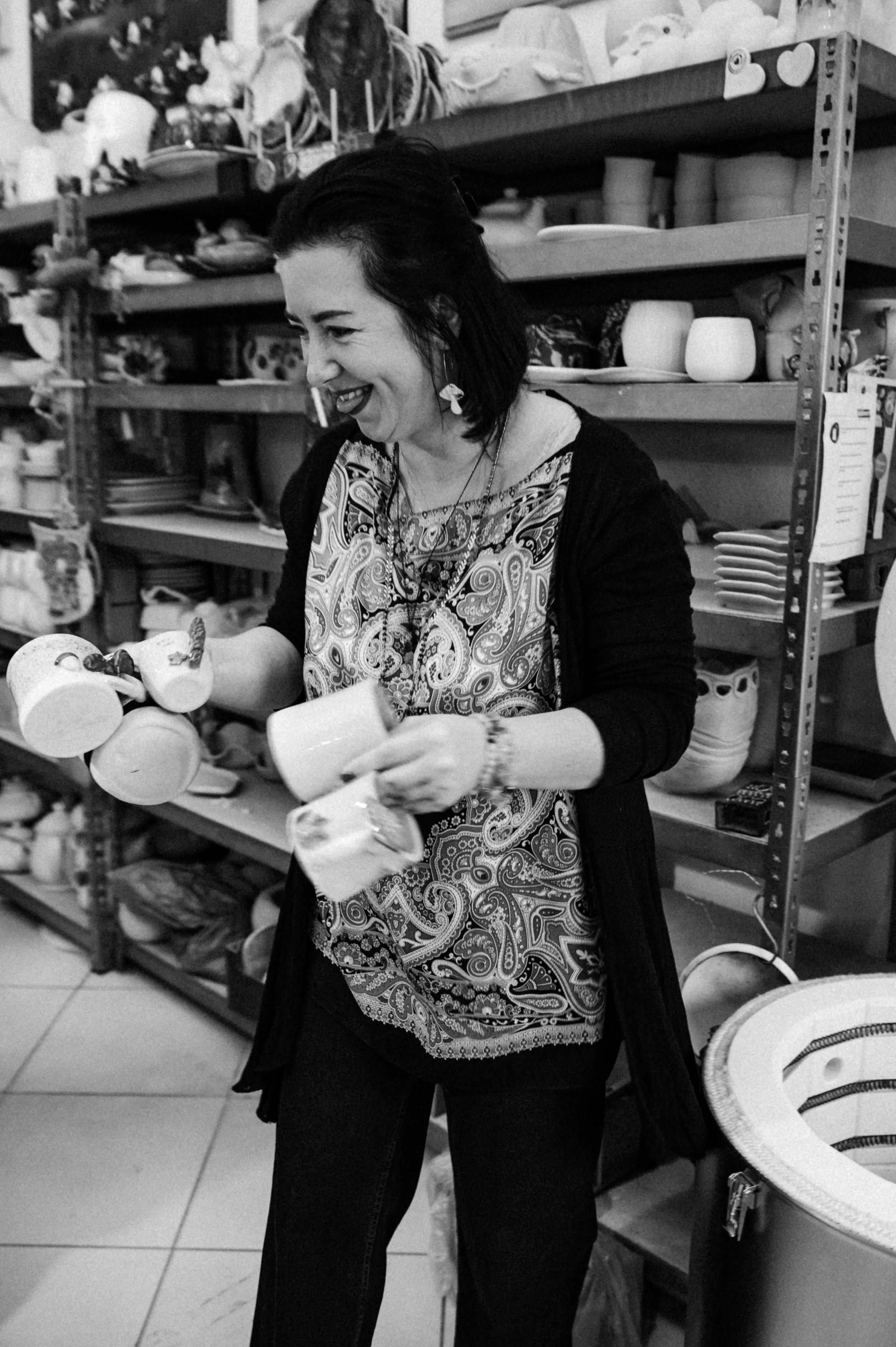 A woman shopping for ceramic mugs at a store, smiling and holding multiple mugs in her hands.