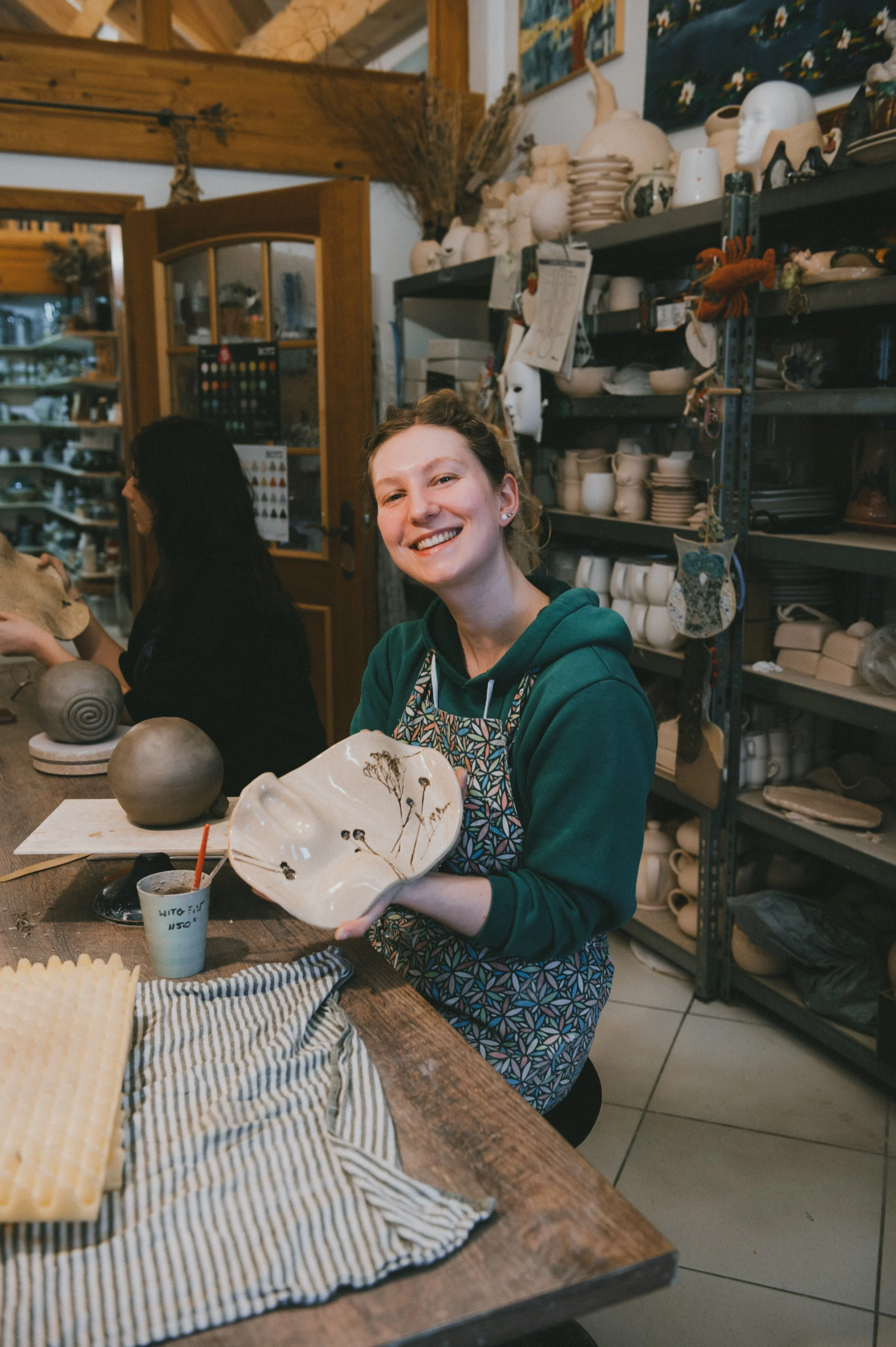 A young woman with a big smile holding a ceramic bowl with a floral design, sitting at a worktable in a pottery studio, surrounded by shelves with ceramics and pottery tools.