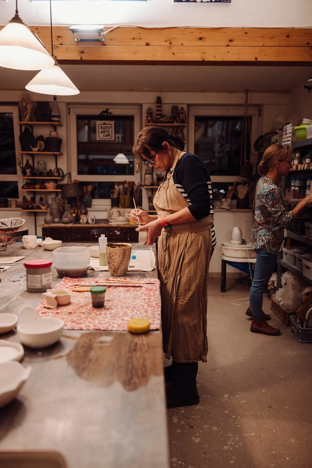 Two women working in a pottery studio; one is painting a ceramic piece, and the other is organizing supplies on shelves.