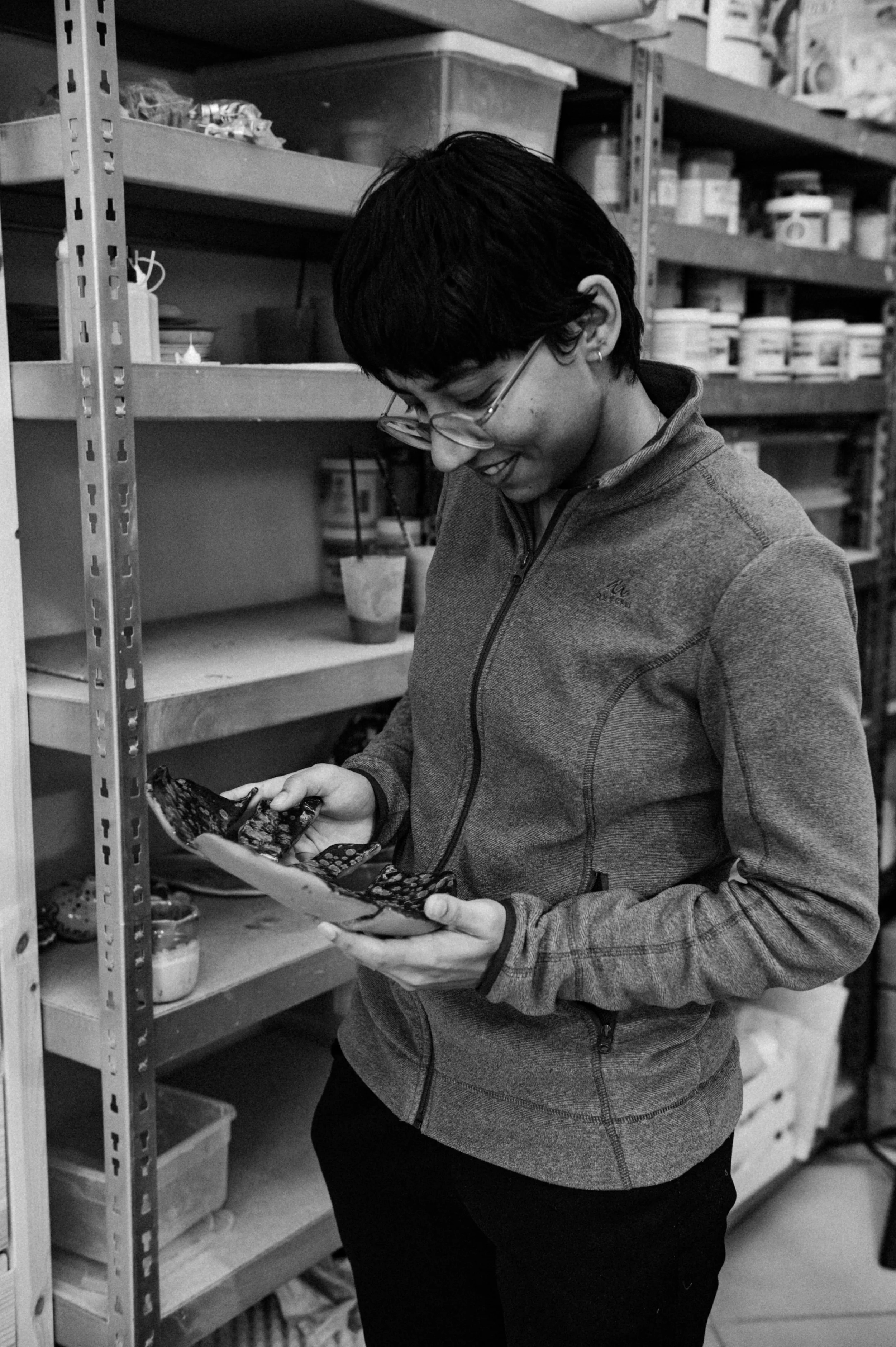 A woman with short dark hair, glasses, and a zip-up jacket smiling while holding and looking at a patterned ceramic bowl in an aisle with shelves filled with containers and jars.
