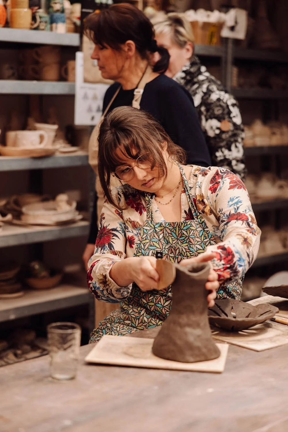 Woman shaping a clay vase in a pottery studio with two women observing in the background.