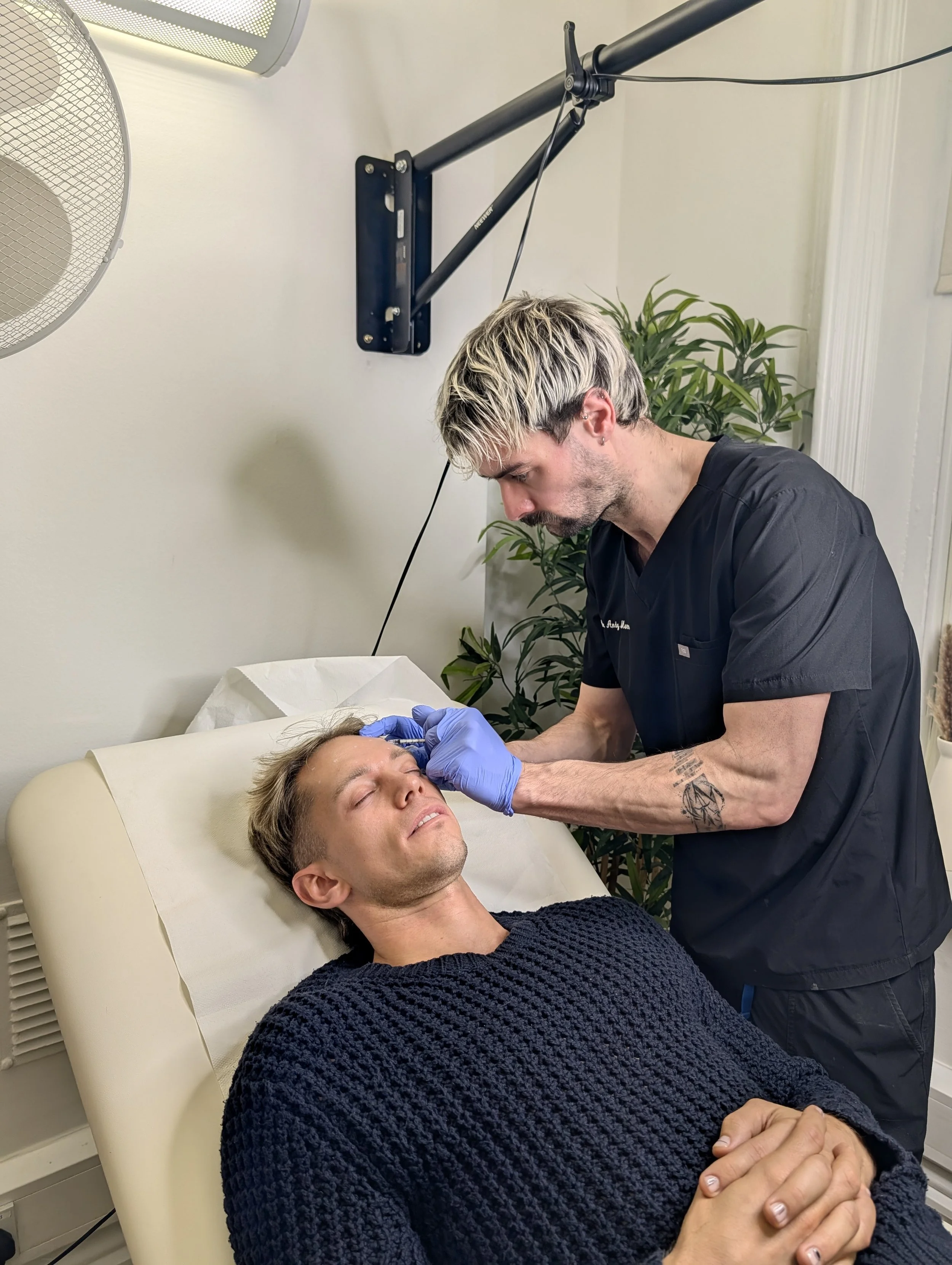 A healthcare professional wearing black scrubs and blue gloves performs a procedure on a man lying on a medical examination bed. The man has his eyes closed and is resting with his hands clasped. There are medical lights and a plant in the background.