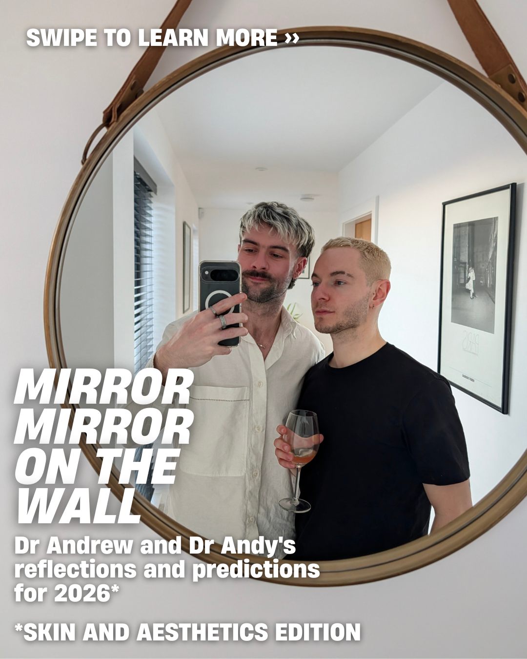 Two men taking a selfie in a mirror, one holding a glass of wine, in a modern room with framed wall art.