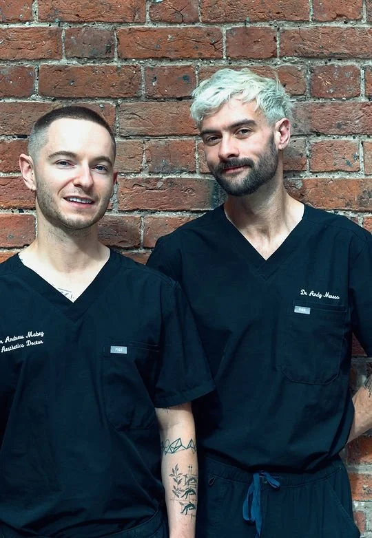 Two male healthcare professionals wearing black scrubs, standing in front of a brick wall.