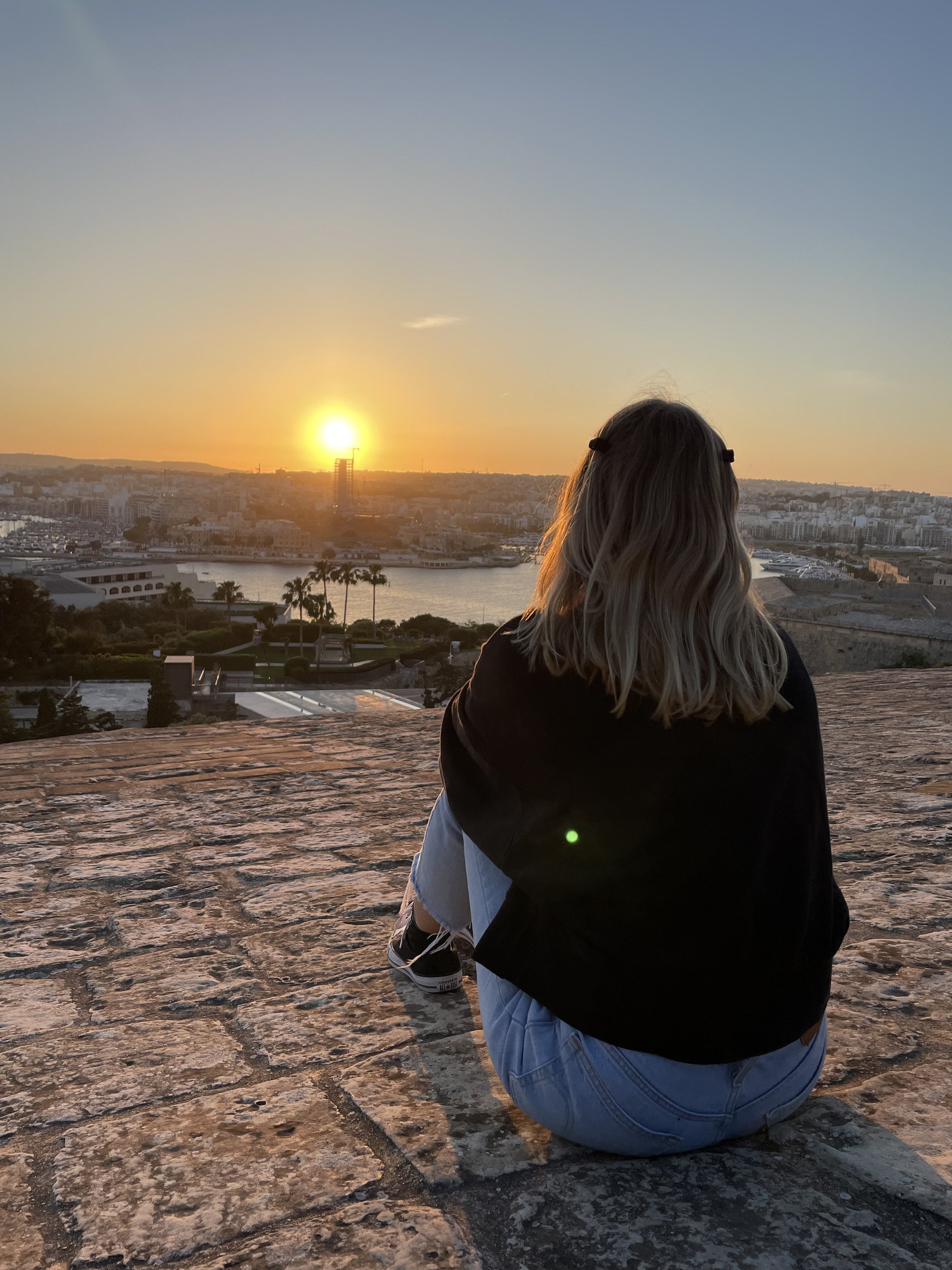Eine Person sitzt auf einer Steinmauer, schaut bei Sonnenuntergang auf eine Wasserstadt.