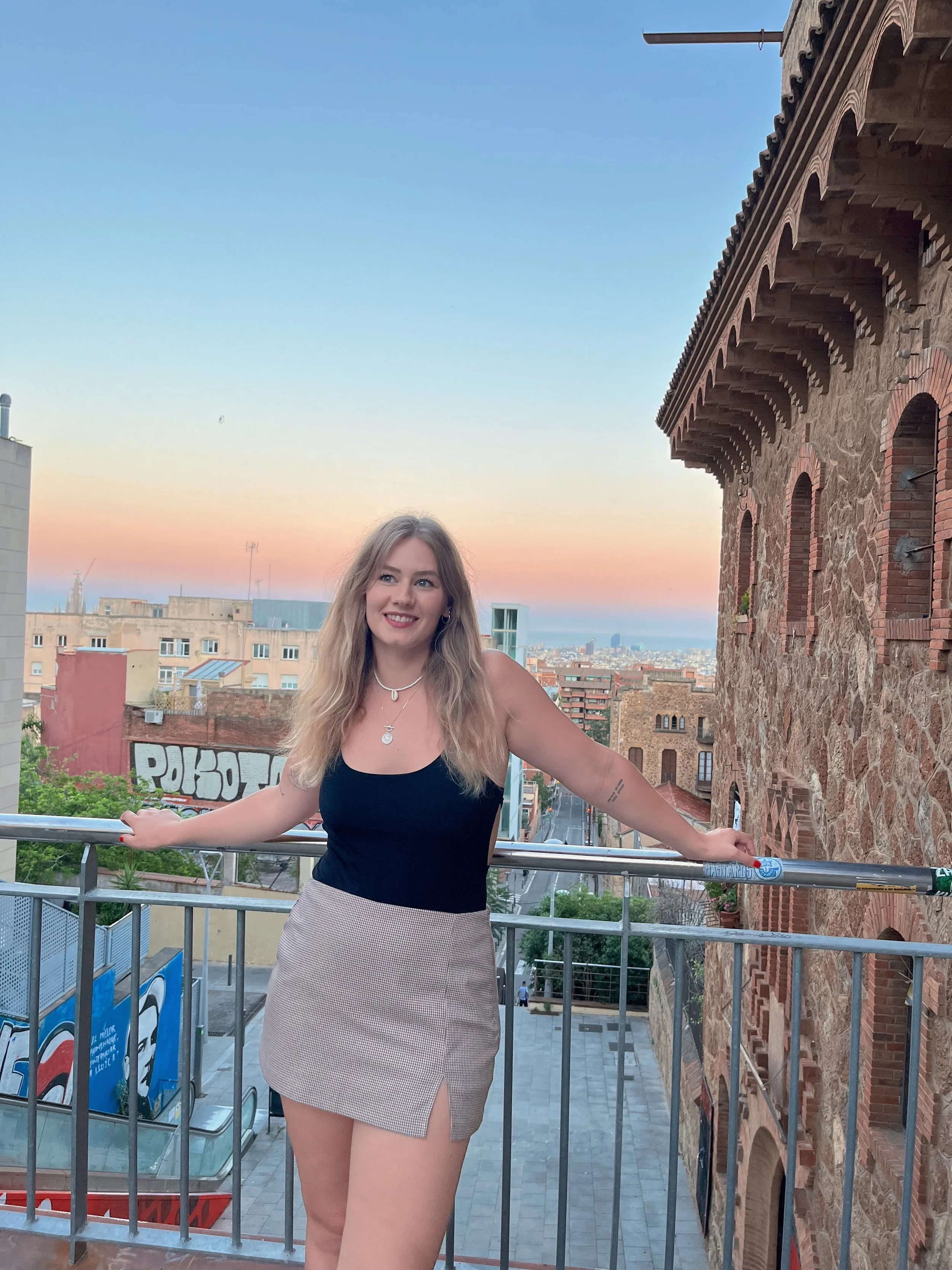 Junge Frau mit blonden Haaren und Gürteltasche, trägt schwarzes Oberteil und beige Rock, steht auf einer Balkonbrüstung vor einer urbanen Stadt im Sonnenuntergang.
