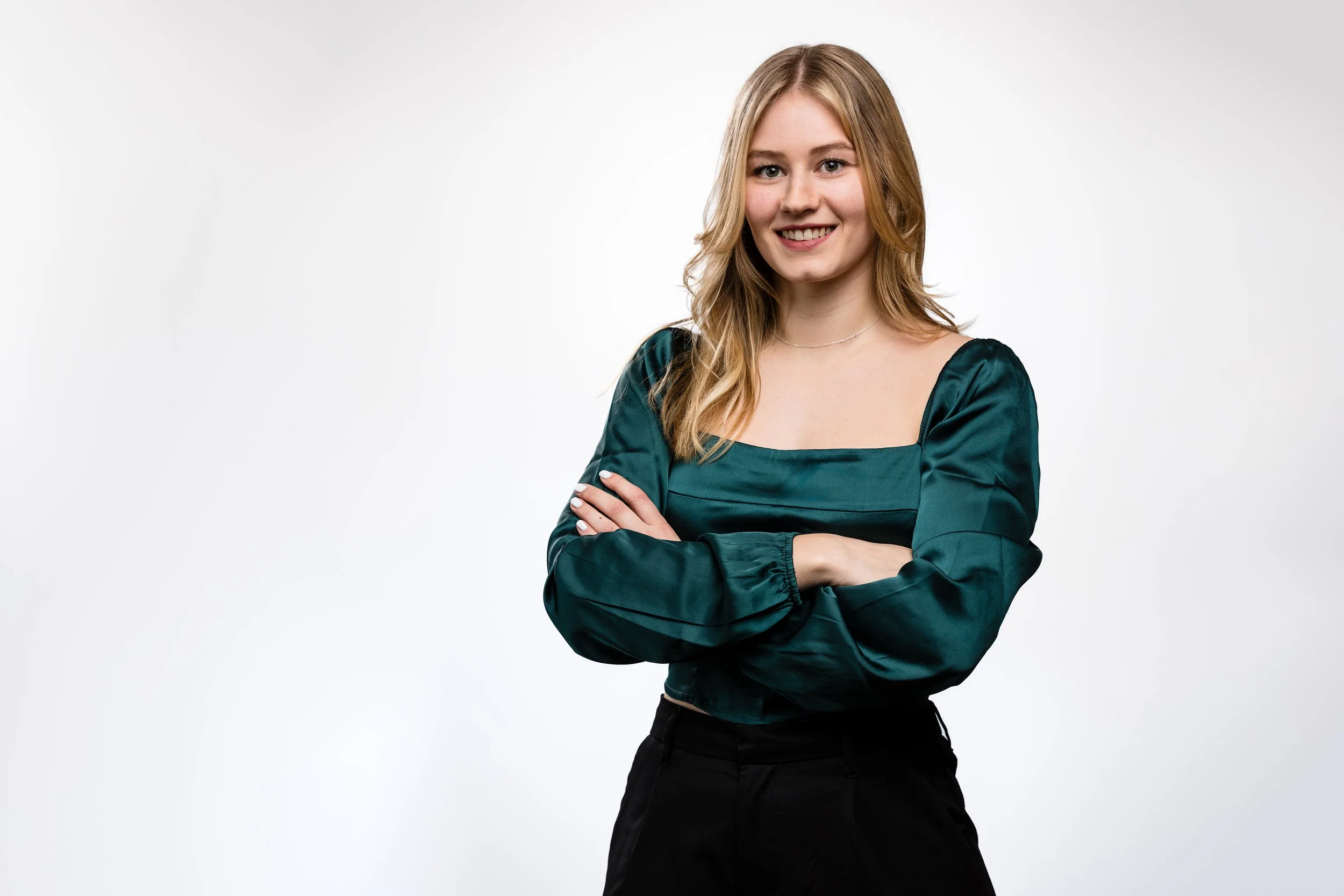 Junge Frau mit blonden Haaren, grünem Satinkleid und schwarzen Hosen posiert vor weißem Hintergrund und lächelt.