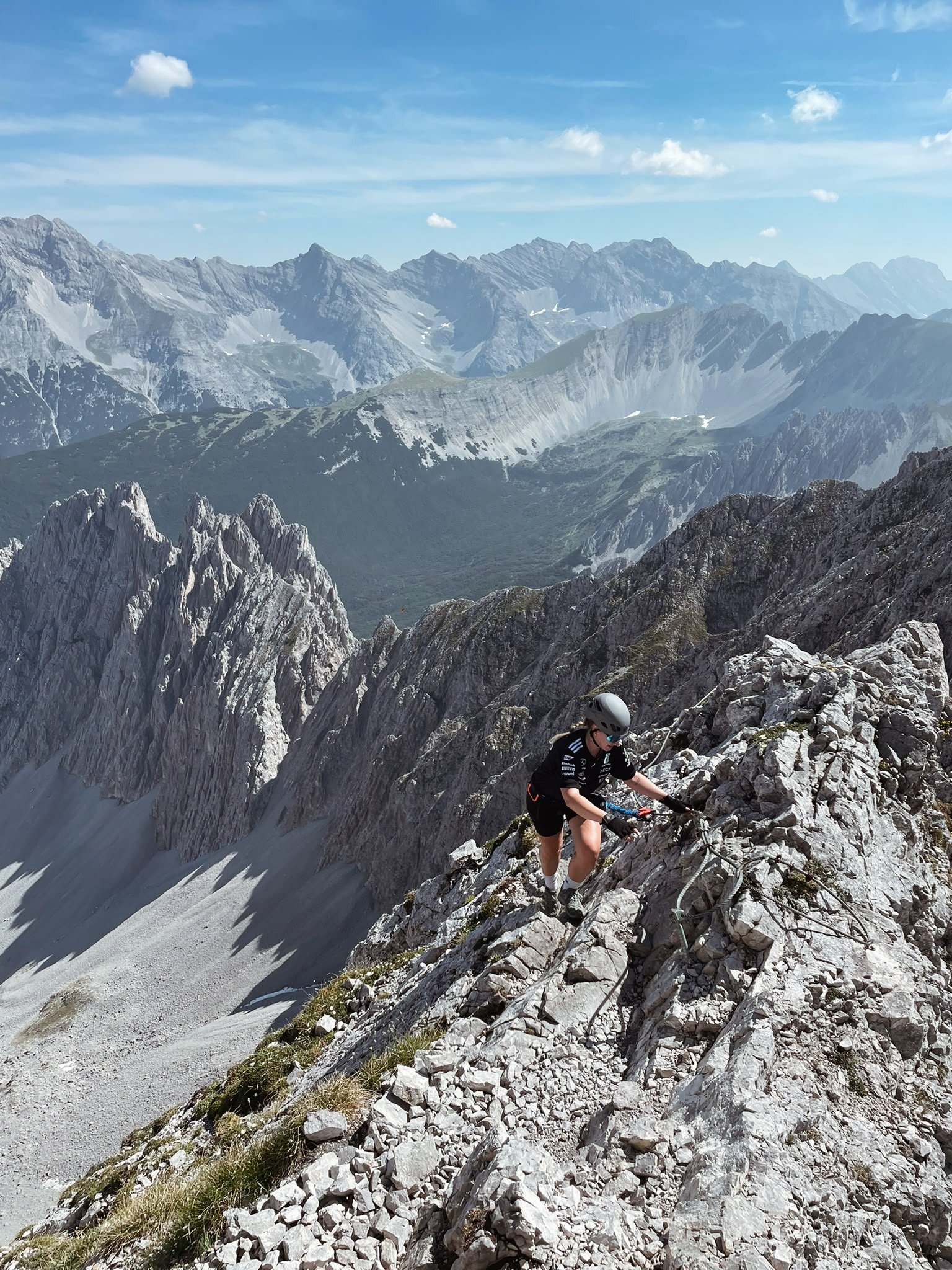 Ein Bergsteiger klettert an einem rutschigen, steilen Berghang mit Felsbrocken und Seilen, umgeben von hohen, schneebedeckten Bergen und einer blauen Himmel mit wenigen Wolken.