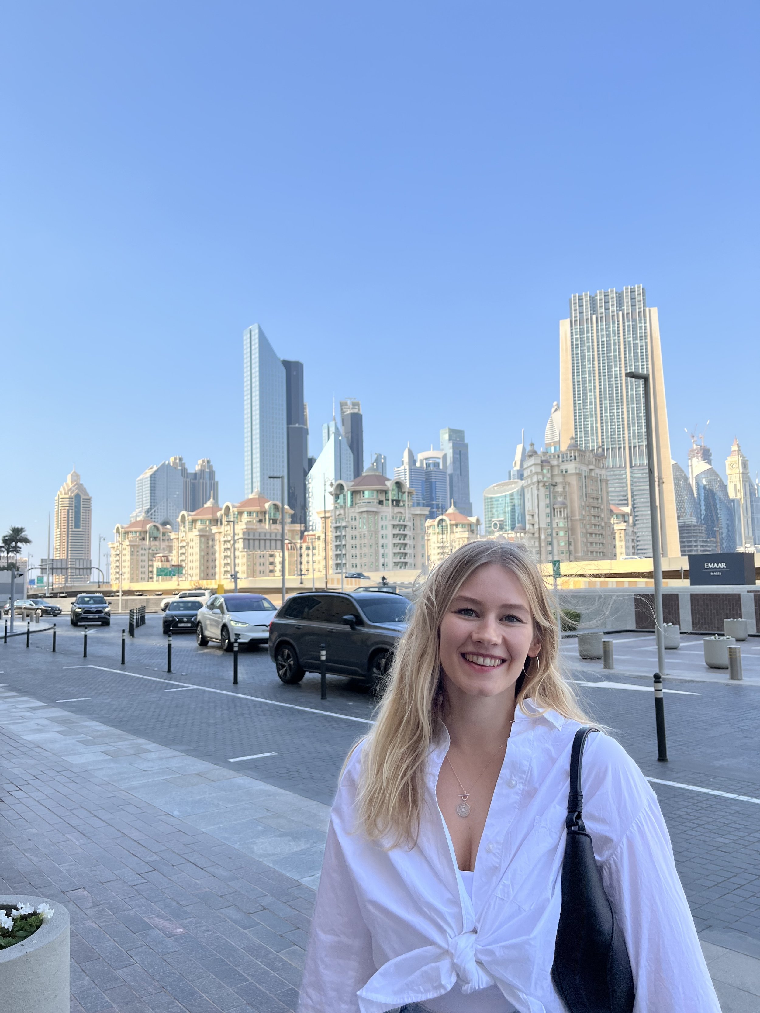 Eine junge Frau mit blonden Haaren und weißem Hemd steht vor einer Skyline mit modernen Hochhäusern in Dubai. Sie lächelt und trägt eine Handschlaufe.