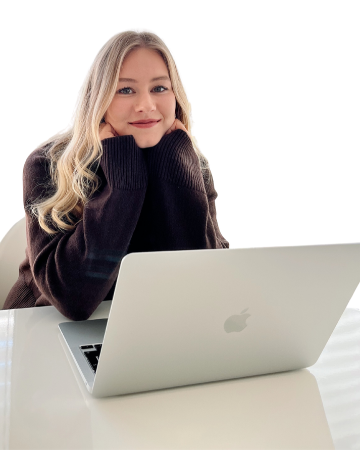 Junge Frau mit blonden Haaren sitzt an einem weißen Tisch mit einem offenen MacBook, trägt einen dunkelbraunen Pullover und lächelt.