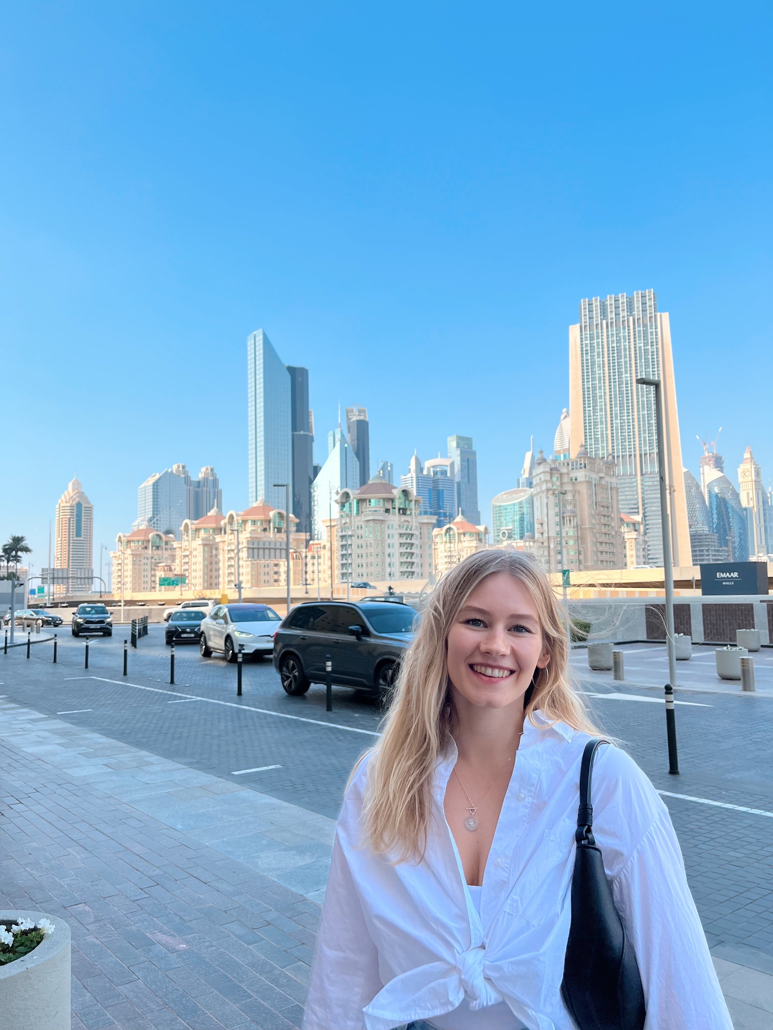 Lächelnde junge Frau mit langem blondem Haar, weißem Hemd und schwarzer Tasche vor einer Skyline mit modernen Hochhäusern und blauer Himmel.