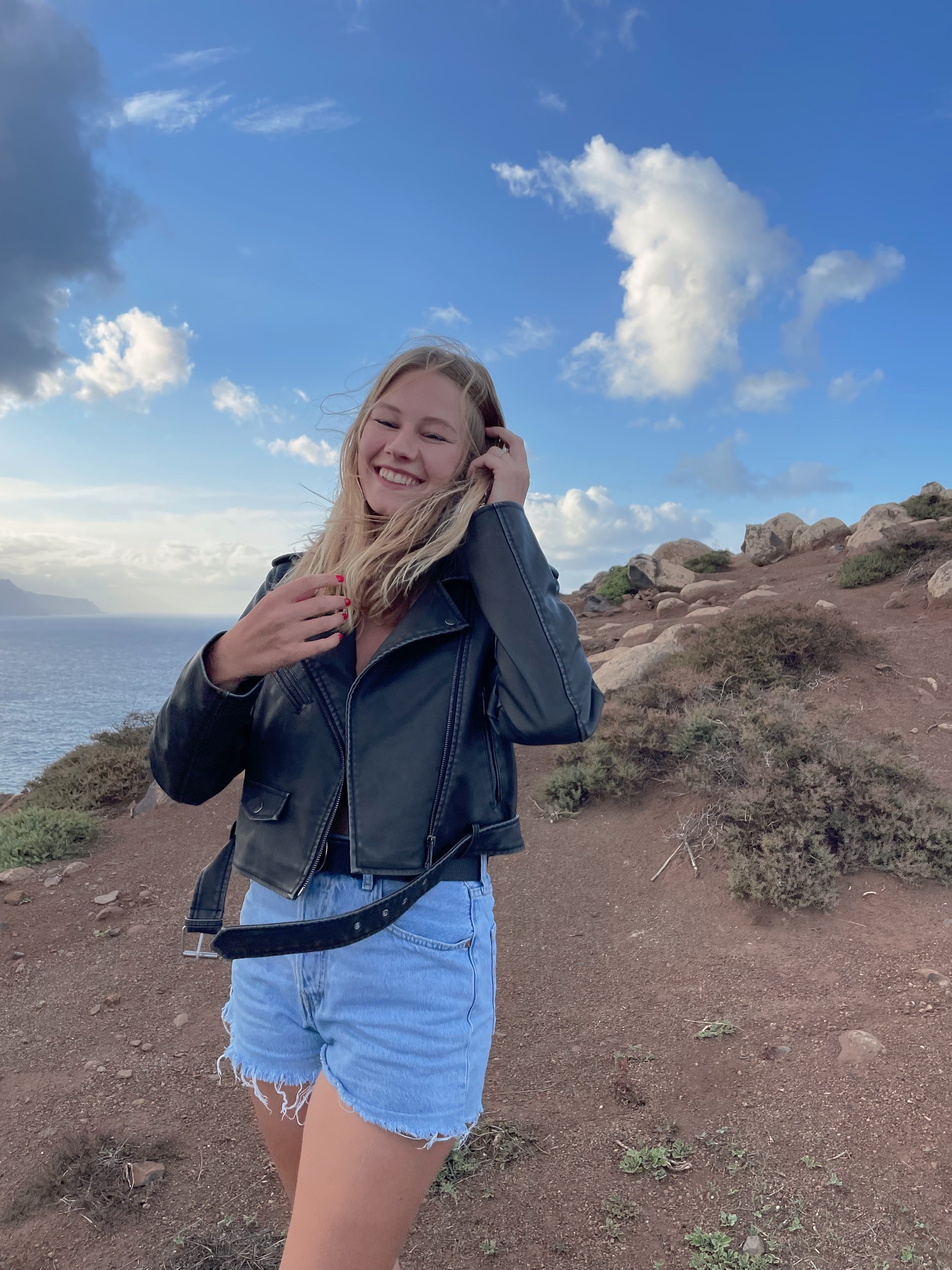 Junge Frau mit lockigem Haar, lachend, trägt eine schwarze Lederjacke und blaue Jeansshorts, steht auf einer felsigen Küstenlandschaft unter einem blauen Himmel mit weißen Wolken.