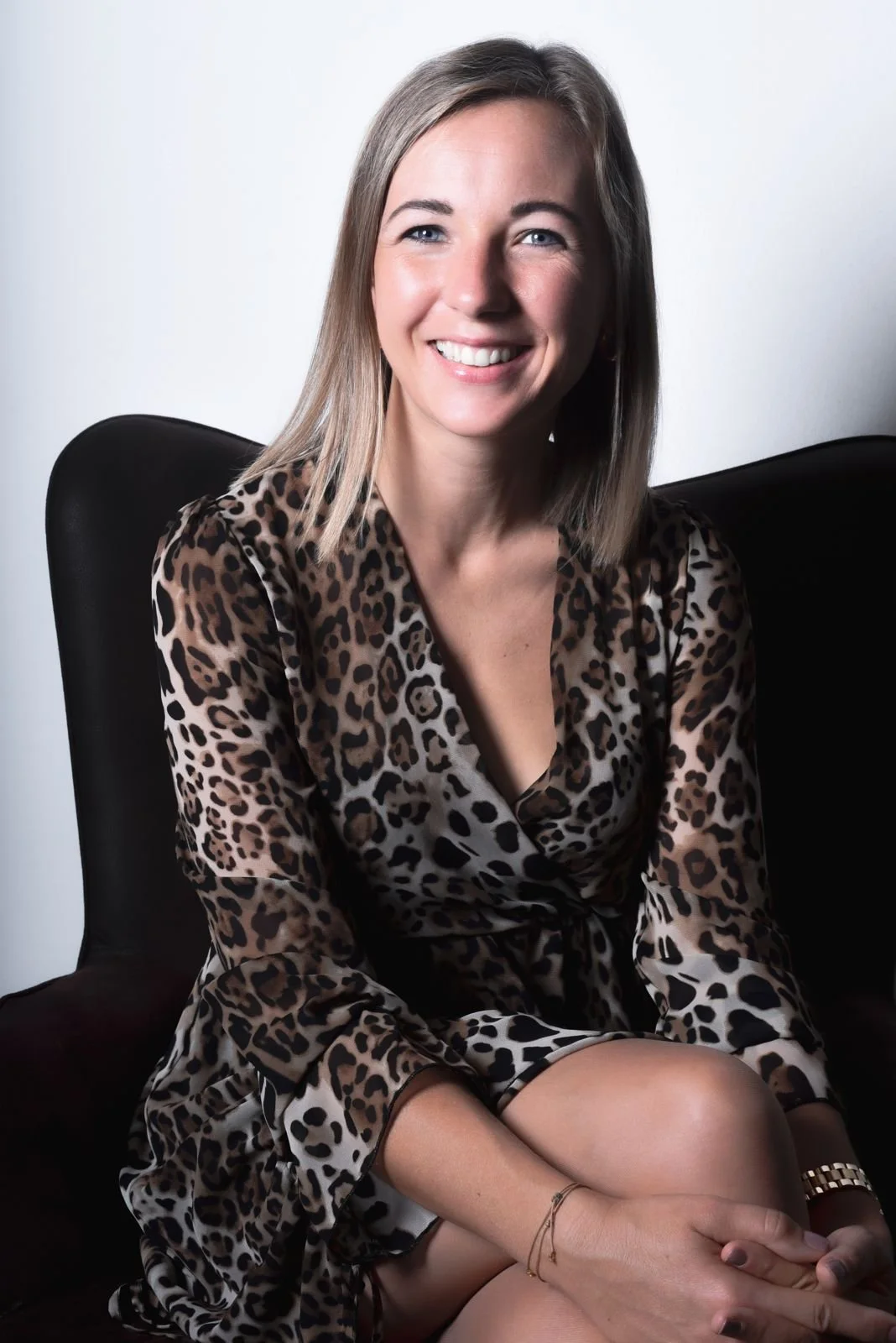 Junge Frau mit blonden, schulterlangen Haaren, lachend, trägt ein Kleid mit Leo-Muster, sitzt auf einem schwarzen Stuhl vor weißem Hintergrund.