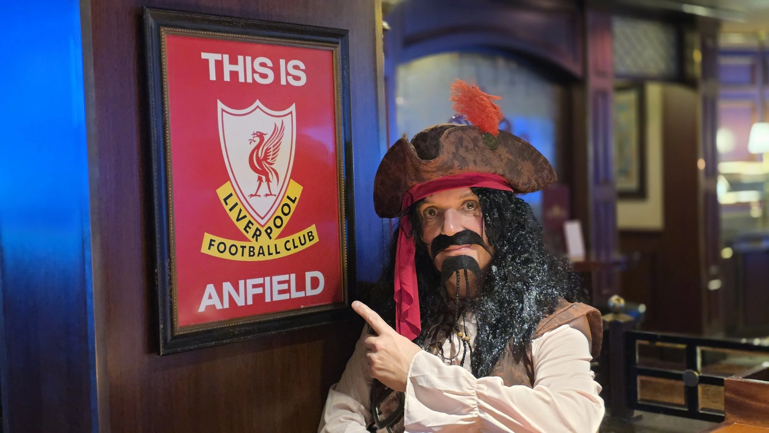 A person in pirate costume, wearing a large brown pirate hat with a red feather, a black curly wig, a mustache and beard, points at a framed Liverpool Football Club 'This is Anfield' sign on a wooden wall inside a pub or restaurant.