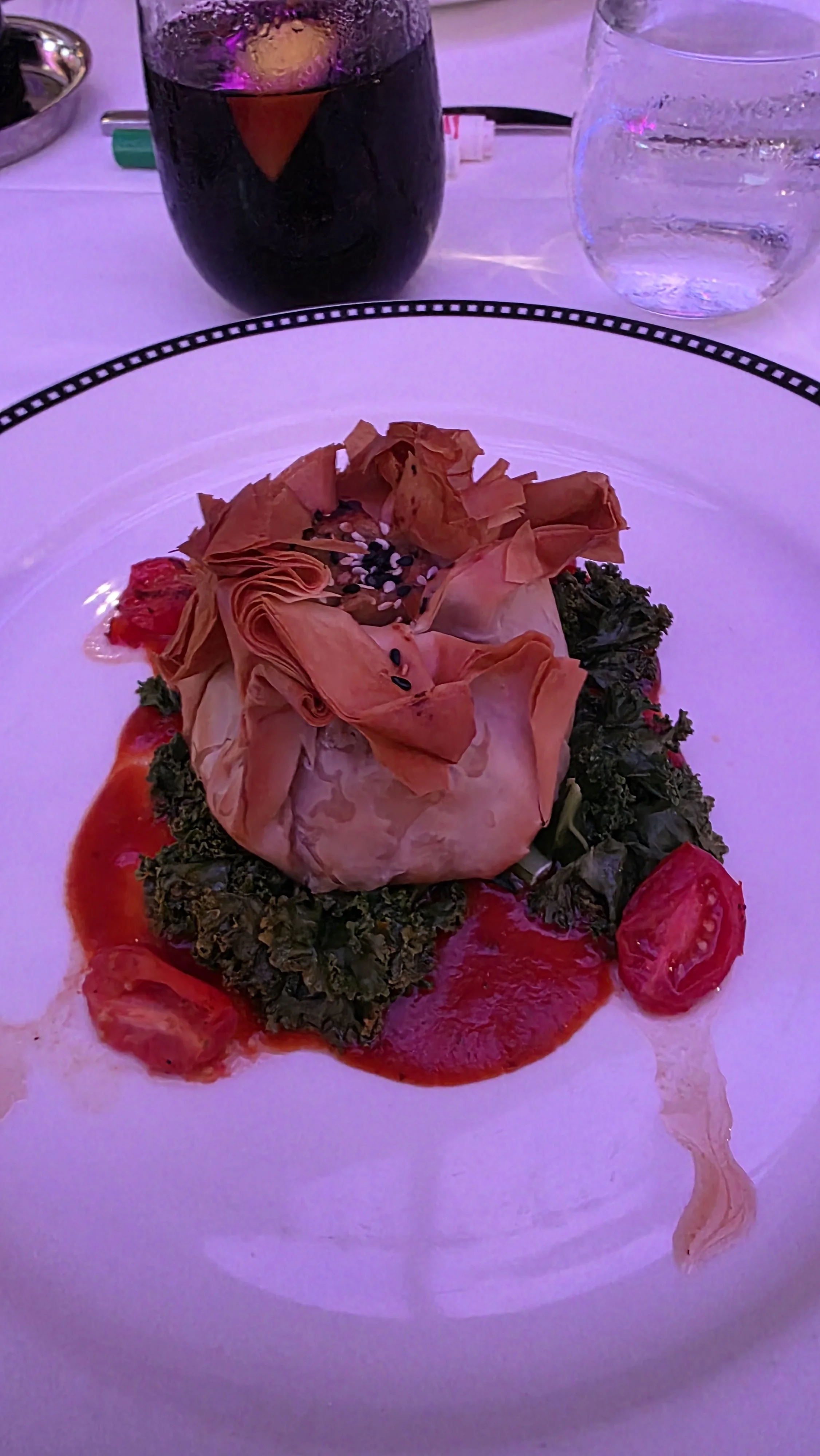 A gourmet dish featuring a puff pastry centerpiece on a bed of leafy greens, with red sauce and cherry tomatoes around it, served on a white plate with a black decorative rim. In the background, there is a glass of red wine, a glass of water, and a w