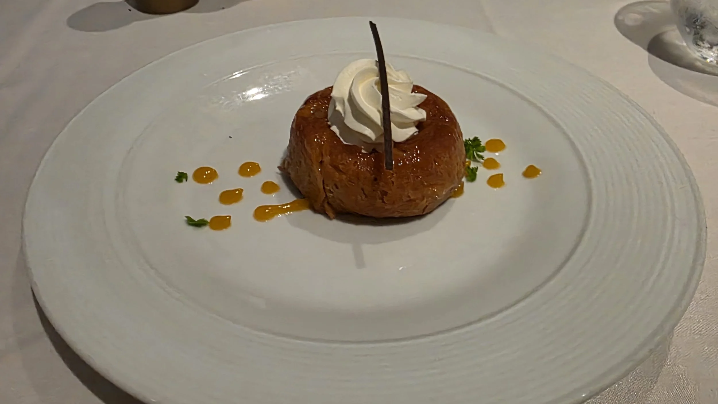 Dessert on a white plate, featuring a caramelized bun with a dollop of whipped cream on top, garnished with a chocolate stick, small green herbs, and orange sauce dots.