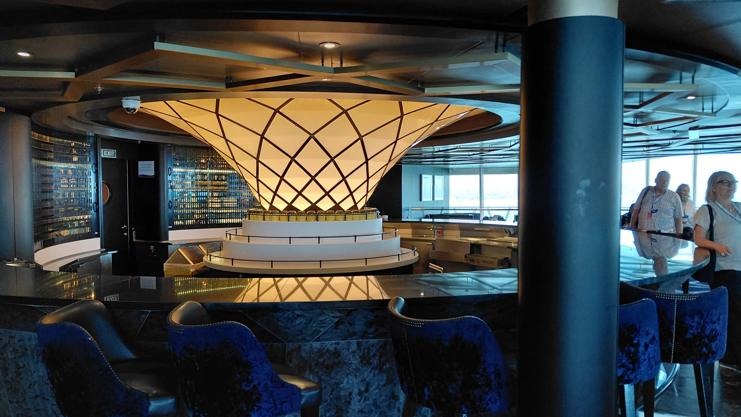 Interior of a modern bar or lounge with blue velvet chairs, a large illuminated cone-shaped light fixture, and a panoramic window view. There are a few people standing near the bar area.