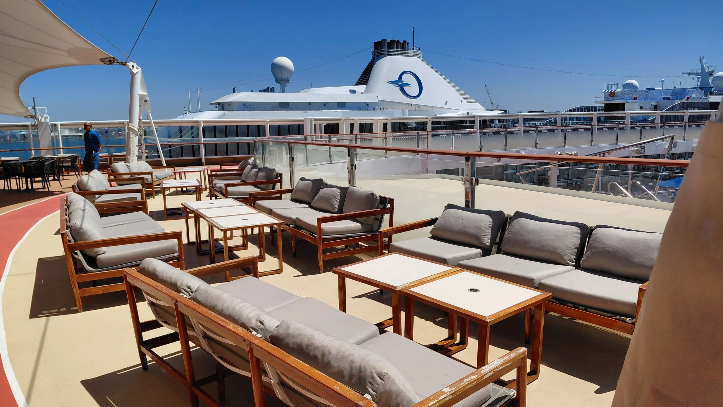 Outdoor lounge area on a cruise ship with wooden framed gray cushioned chairs and tables, overlooking a docked large white cruise ship with a blue logo, under a clear blue sky.