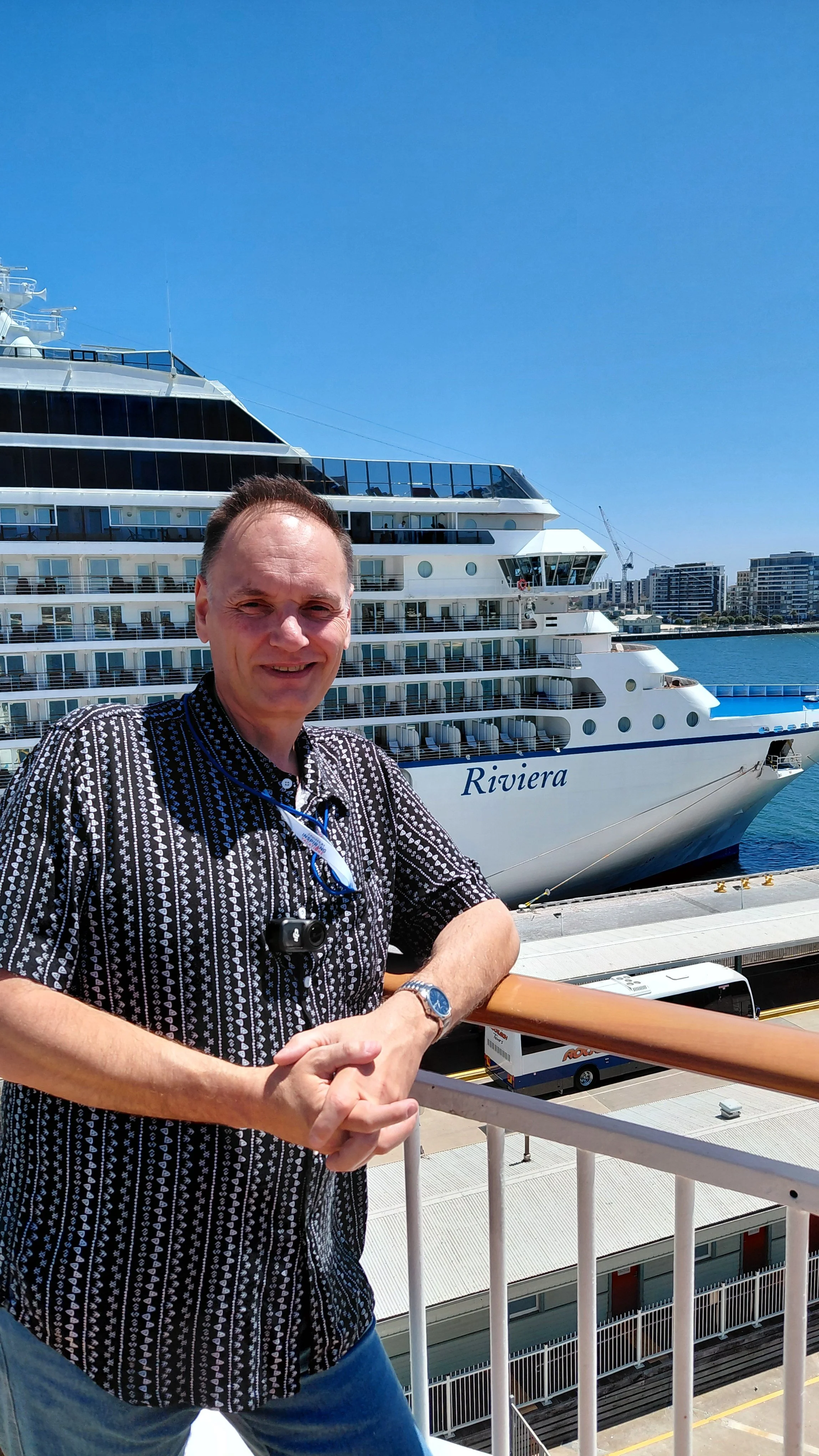 A man standing on a ship's deck with a large cruise ship named Riviera in the background, docks and city buildings visible under a clear blue sky.
