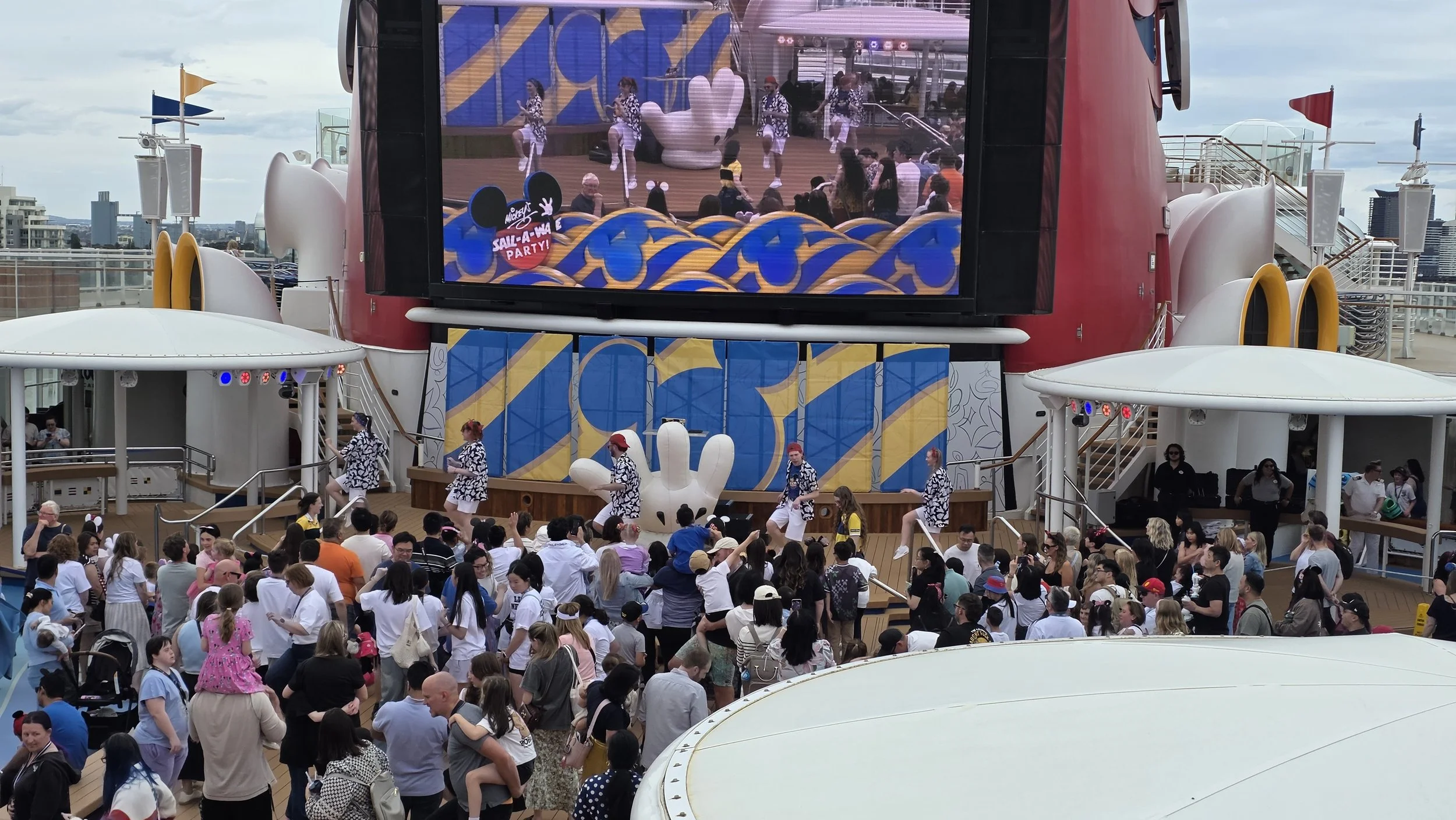 A crowded outdoor event on a cruise ship deck featuring a performance stage with dancers, a large video screen, and Disney-themed decorations and characters, including Mickey Mouse ears and a giant hand.