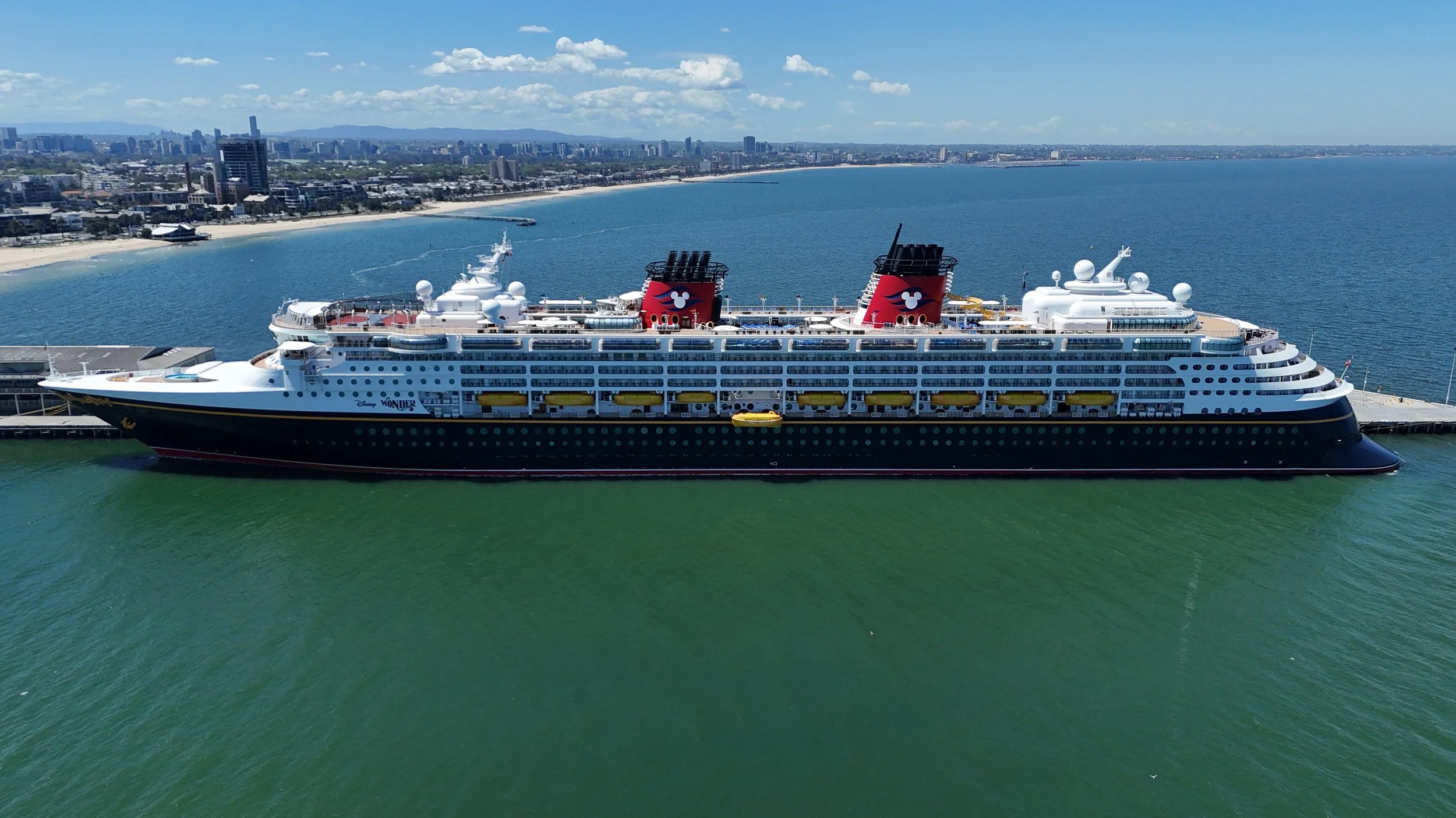 A Disney cruise ship docked at a port with a city skyline and beach in the background on a sunny day.