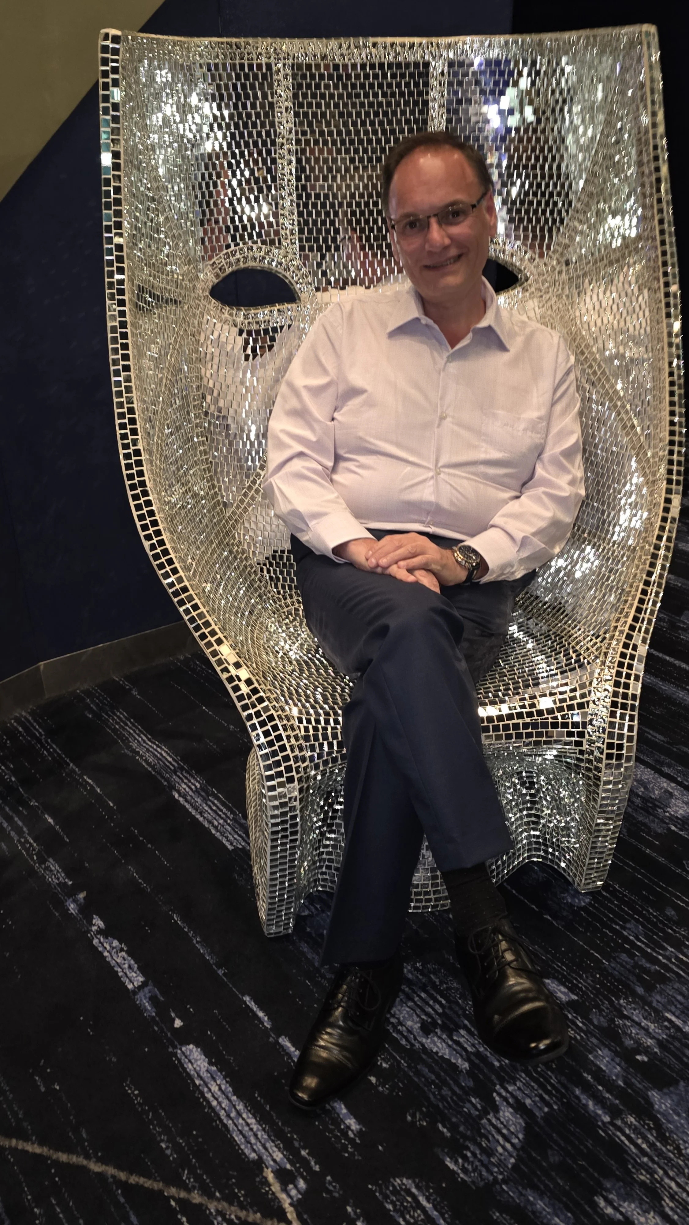A man sitting on a futuristic, reflective, mirrored chair in a dark room.