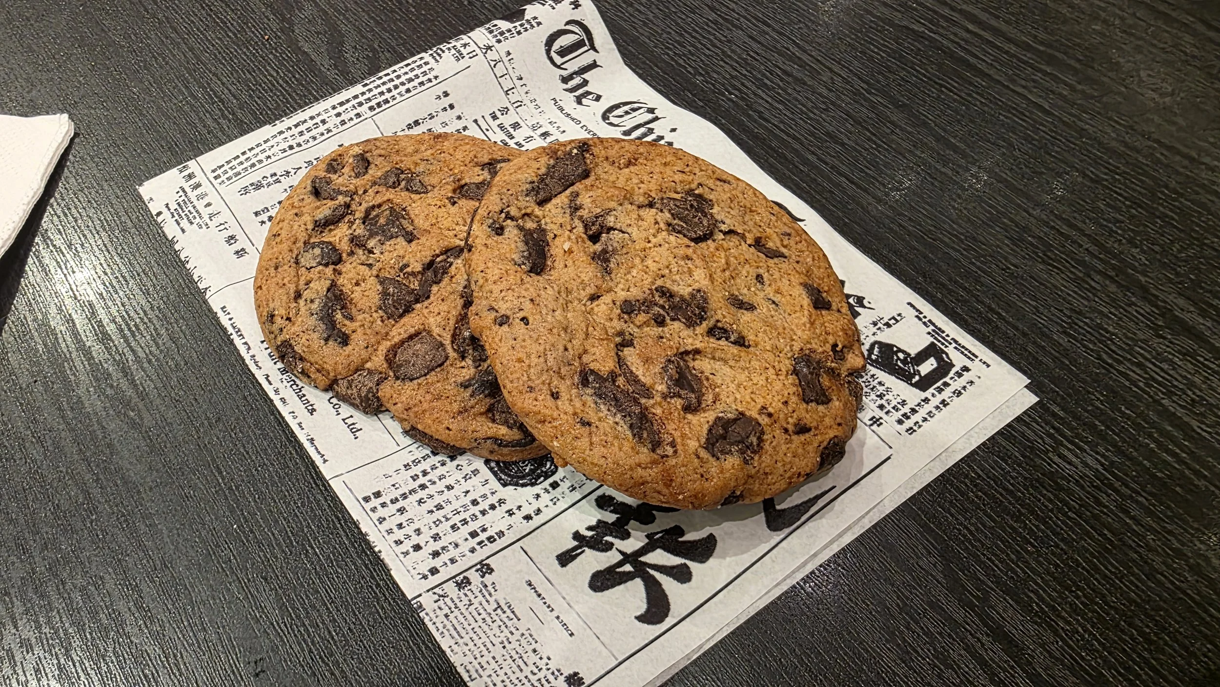 Two chocolate chip cookies on a newspaper on a dark wooden table.